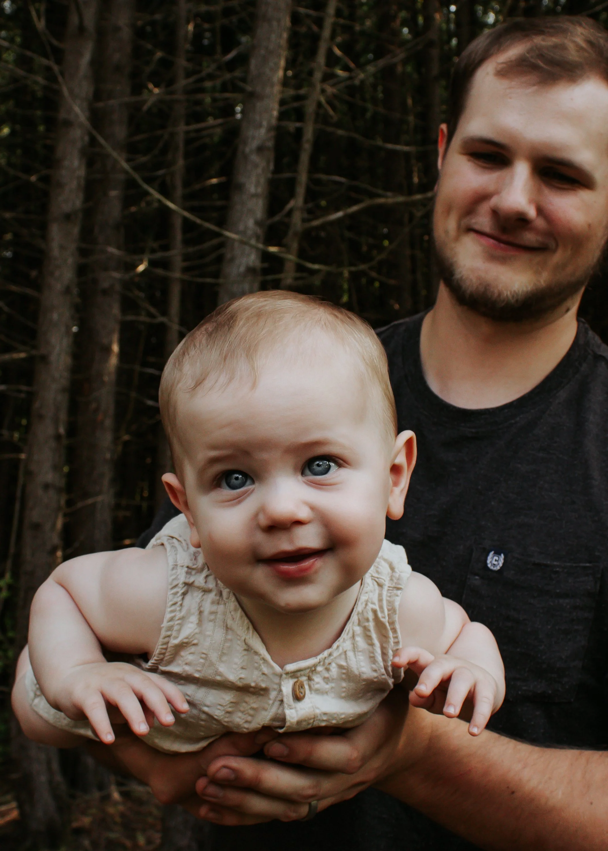 A man holding a smiling baby with blue eyes outdoors, with trees in the background.