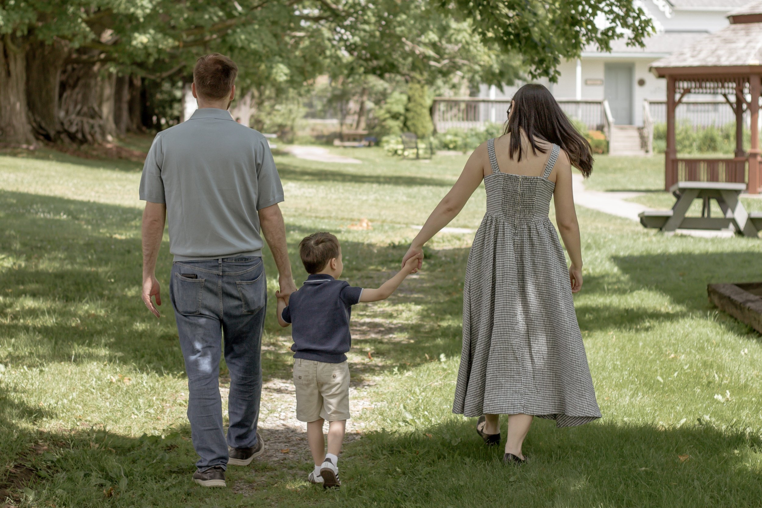 Family of four walking on a path in a park with trees, a gazebo, and a house in the background. Family photos at the uxbridge historical centre