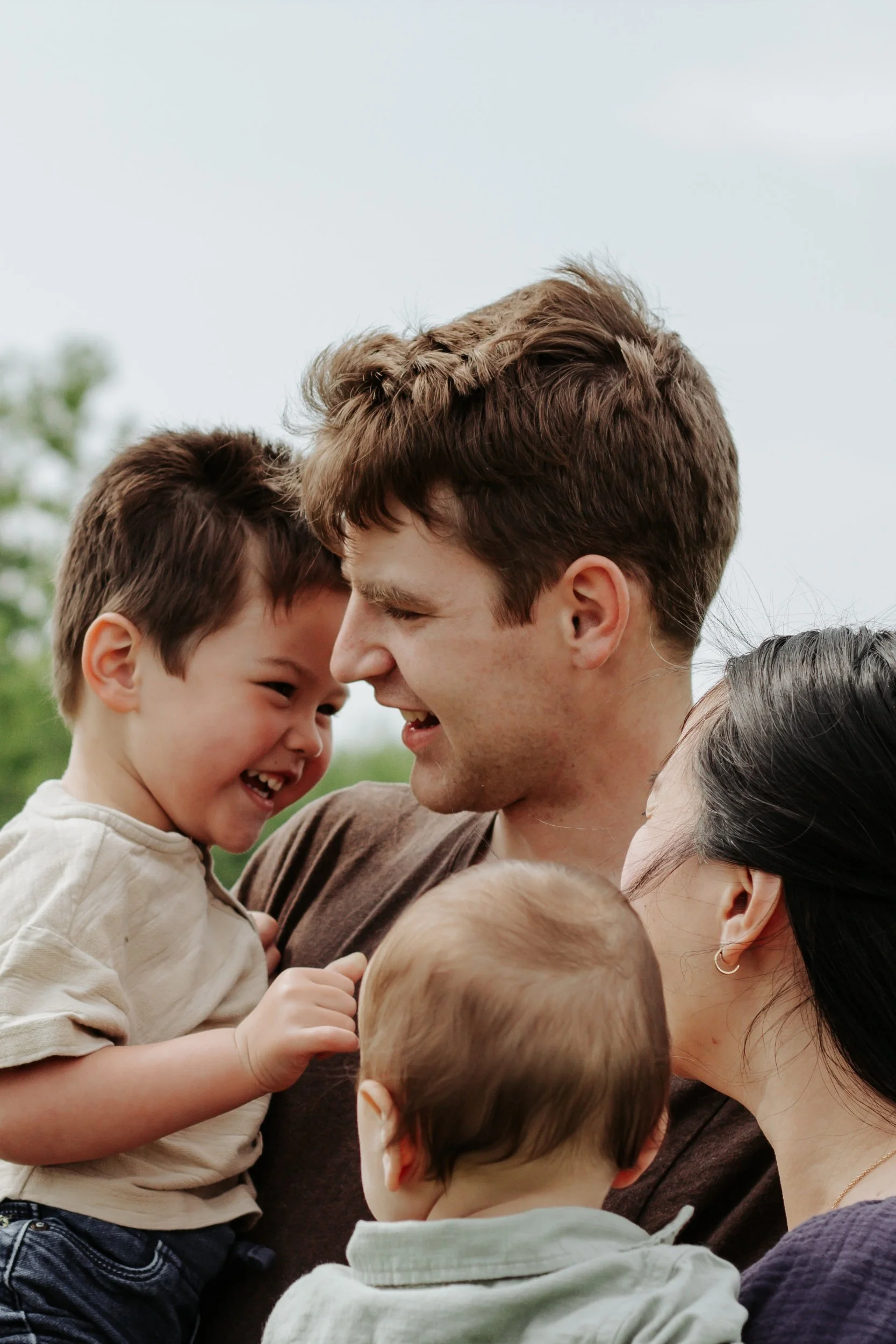 A happy family outdoors, with a father, mother, and two young children, sharing a joyful moment. The father and son are smiling and touching foreheads, while the mother and baby are close together.
