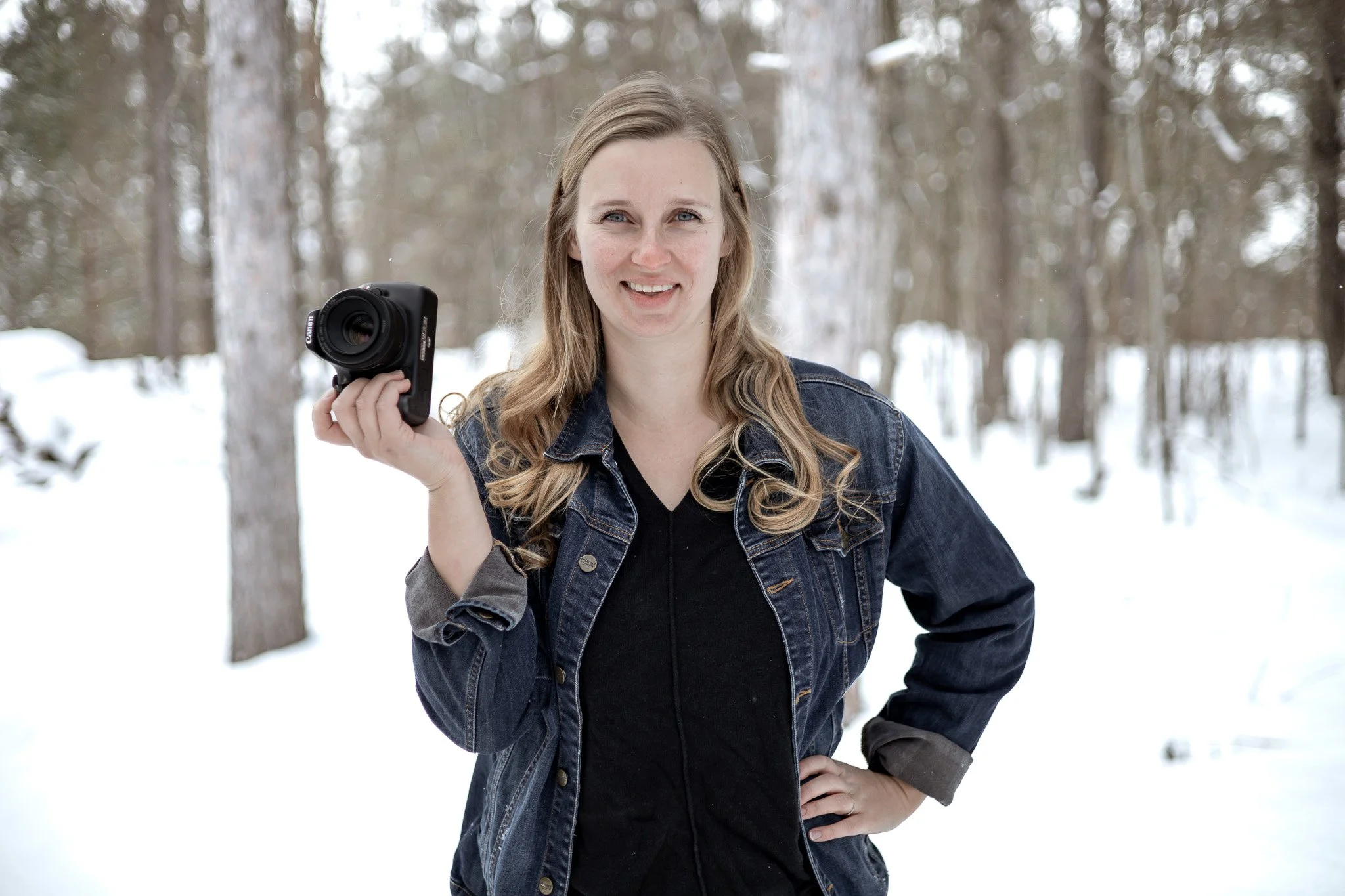 Uxbridge family photographer, Micaela Bouwmeister of Micaela B Photography poses  outdoors in a snowy forest, holding a camera in her right hand, smiling at the camera.