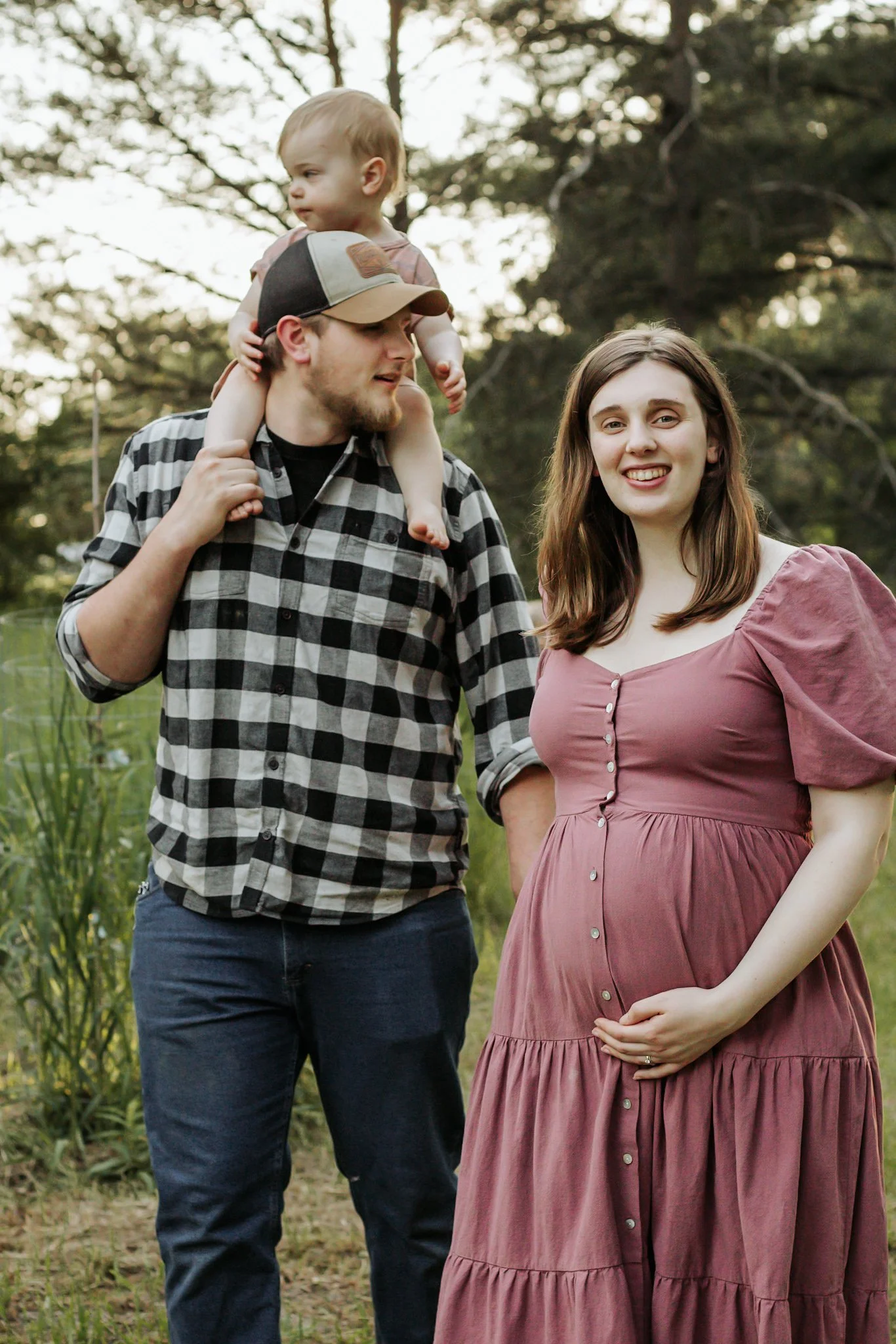 A man carrying a young child on his shoulders stands outdoors with a woman in front of him. The woman is smiling and has one hand on her pregnant belly. The scene is set in a natural environment with trees and greenery. maternity photos Uxbridge onta