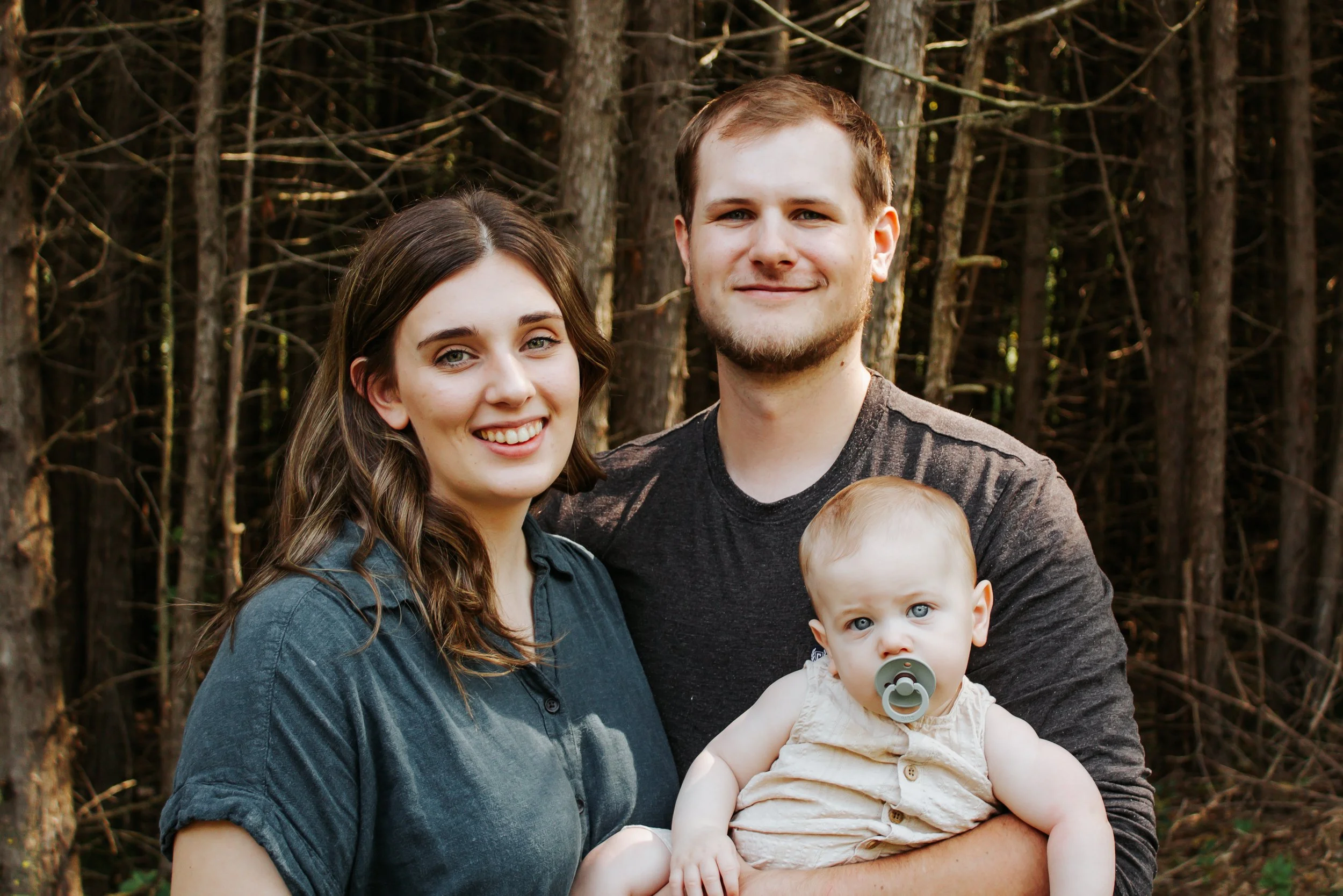 A family of three people standing outdoors in a wooded area, smiling at the camera. The woman has brown hair and is wearing a dark blue shirt. The man has short brown hair and is wearing a dark gray shirt. He is holding a baby with blue eyes, light h