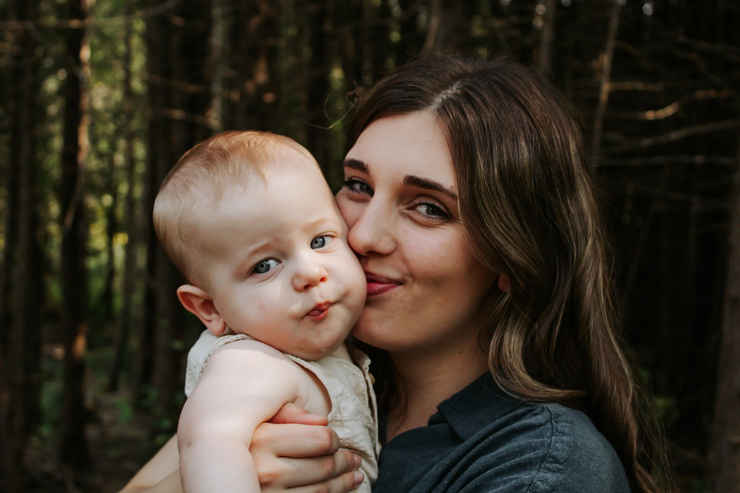 A young woman with brown wavy hair holding a baby with light hair and blue eyes, both outside in a wooded area.