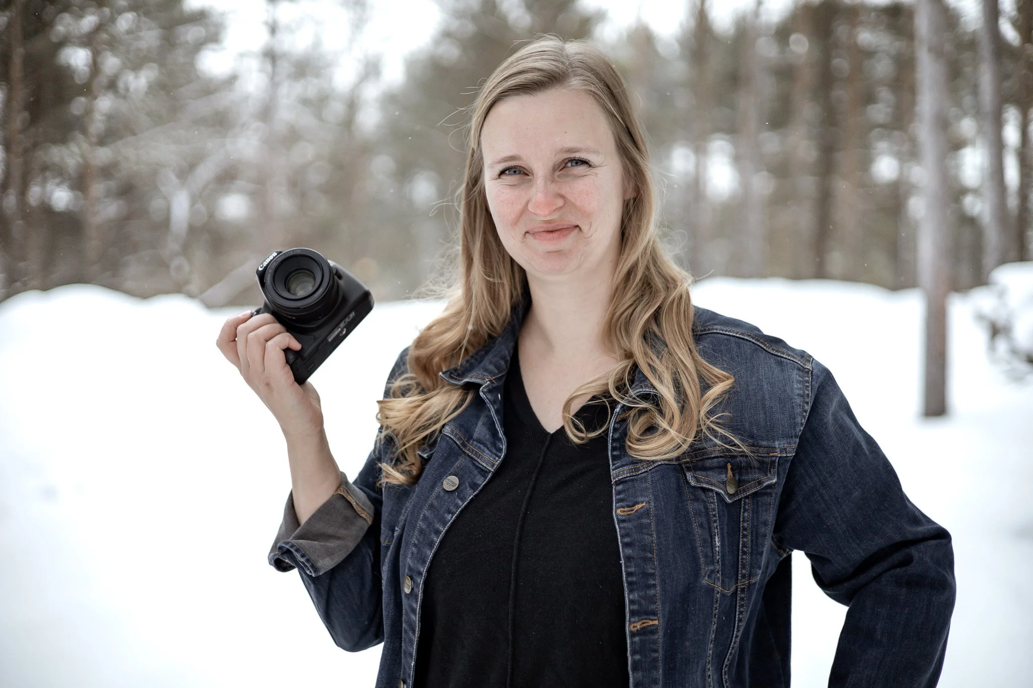 Uxbridge family photographer, Micaela Bouwmeister of Micaela B Photography poses outdoors in a snowy landscape, holding a camera in her right hand, wearing a denim jacket and black shirt.