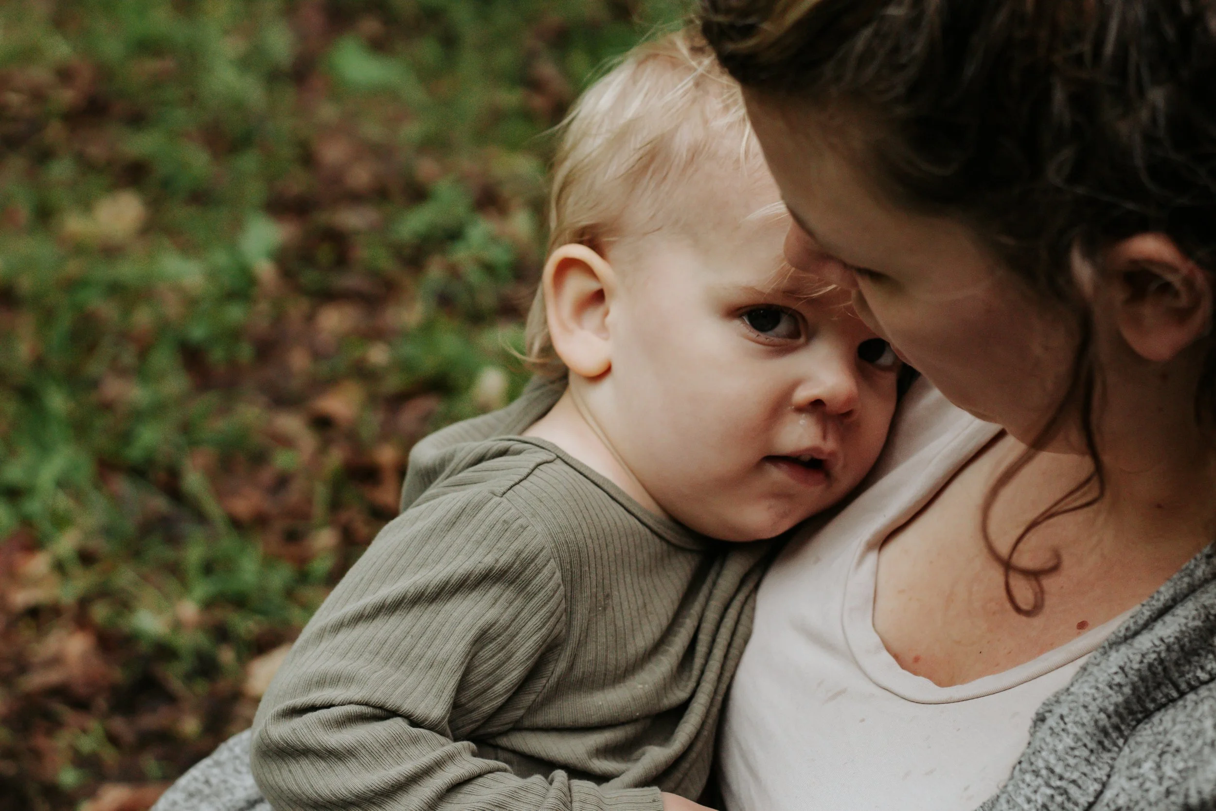 A woman holding a young child outdoors, with the woman gently touching her forehead to the child's, in a close-up shot amid a background of green leaves. Family photos at elgin park uxbridge ontario