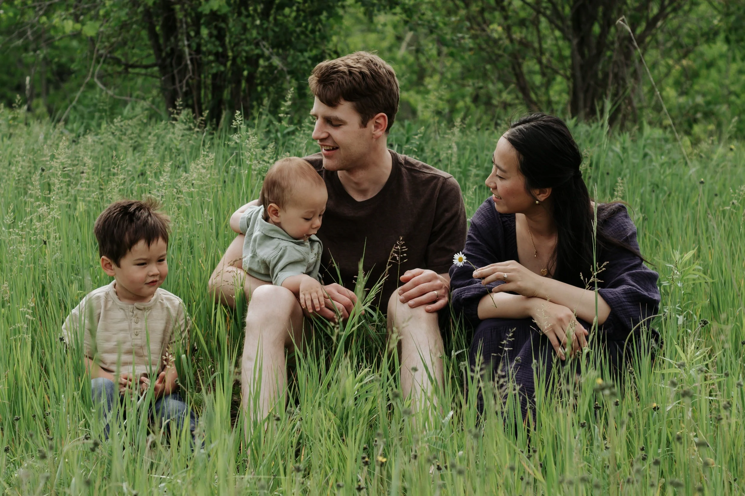 A family of four sitting and playing in a lush green field with trees in the background. Family photos in sunderland ontario