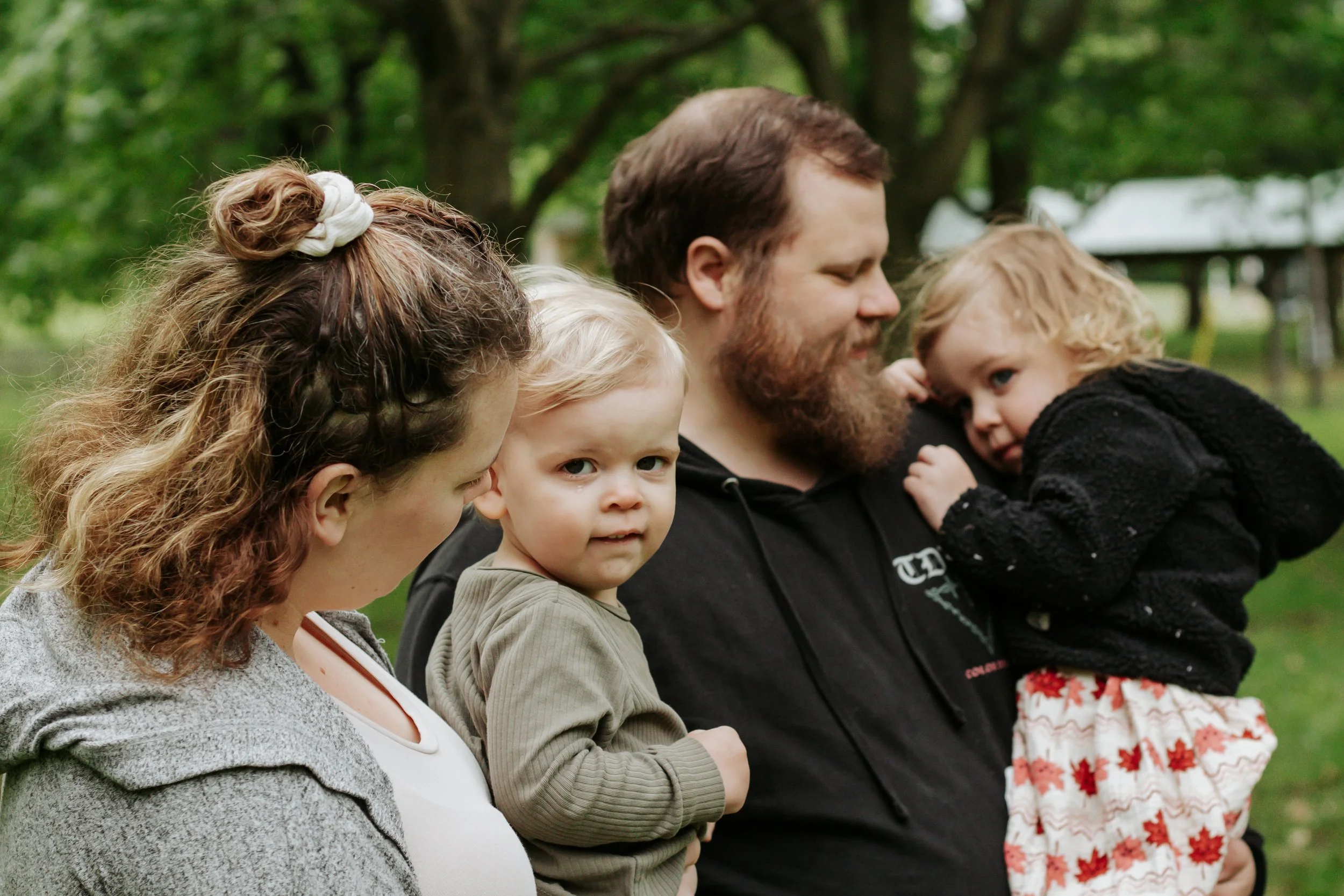 Family of four outdoors, with a woman, a young boy, a man, and a young girl, in a park with trees and a pavilion in the background. Family photos at elgin park uxbridge ontario