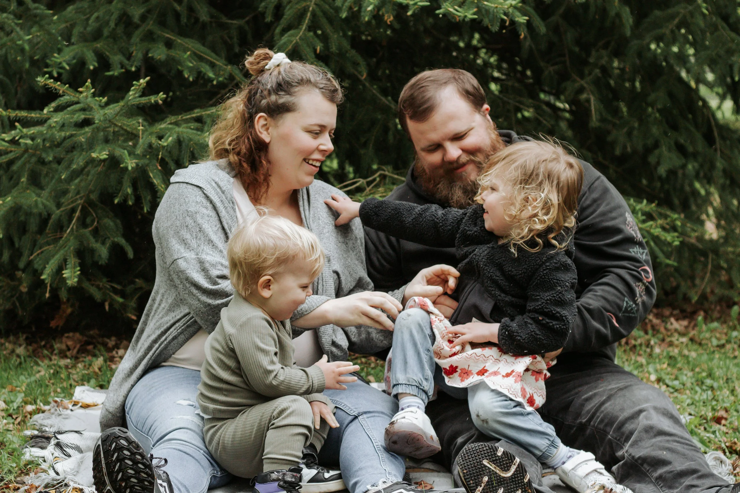 A family of four sitting on a blanket outdoors, surrounded by trees. The mother, father, and two young children are smiling and playing together. Family photos at elgin park uxbridge ontario
