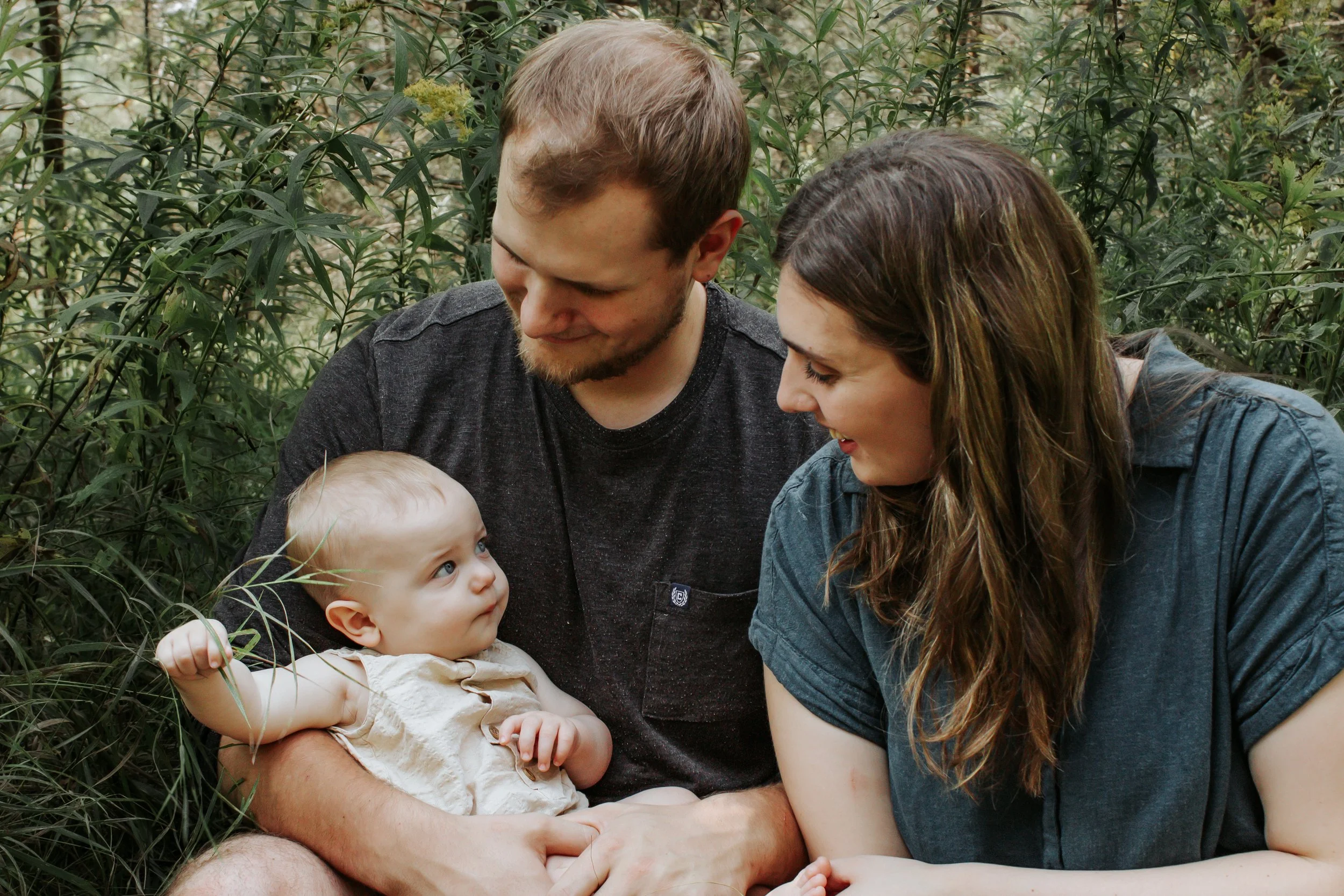 A young family of three outdoors, sitting among green bushes, with a smiling baby girl in the father's arms, looking up at the parents with curious eyes.