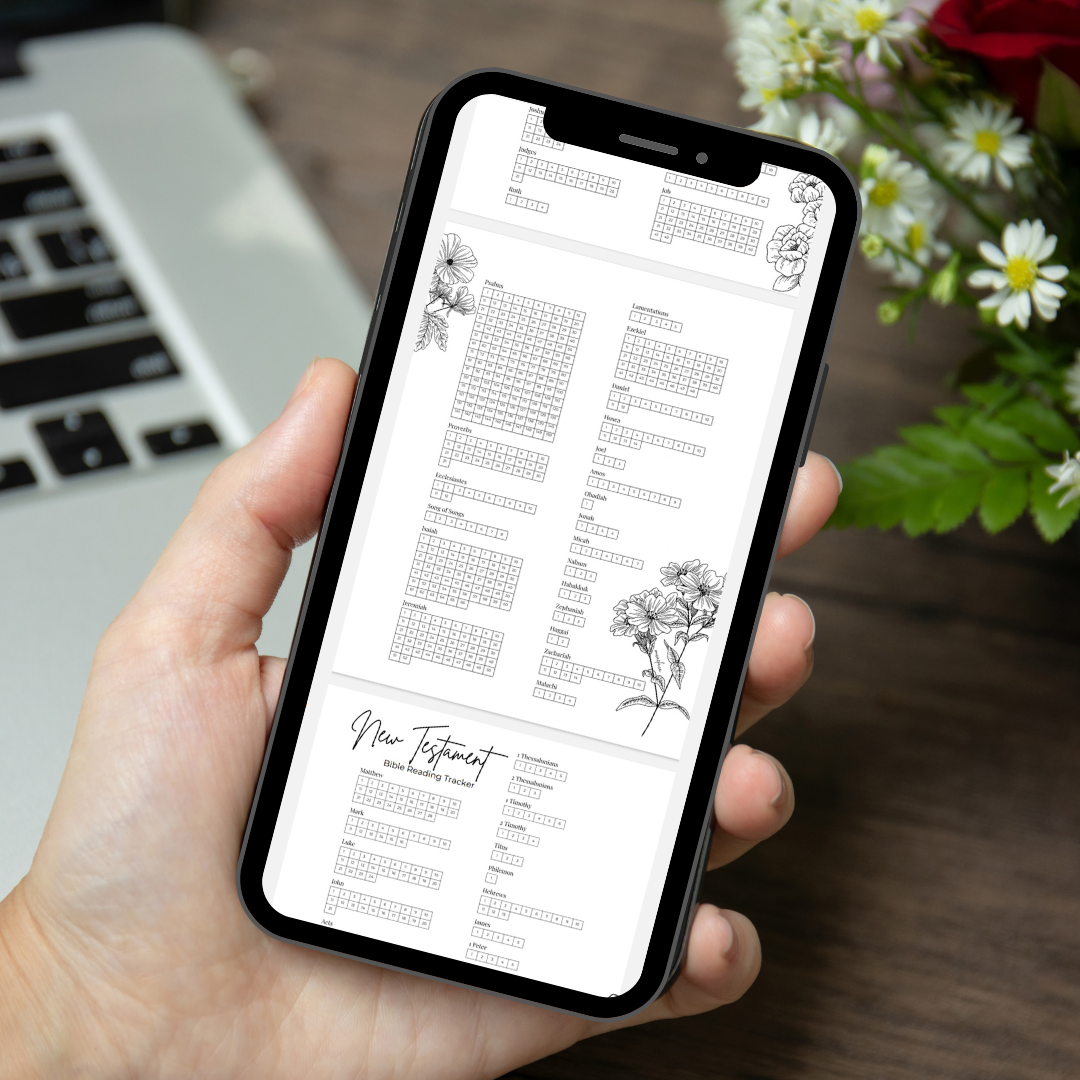 A person holding a smartphone displaying a Bible reading tracker app with various reading schedules and floral illustrations, on a wooden surface next to a laptop keyboard and flowers.