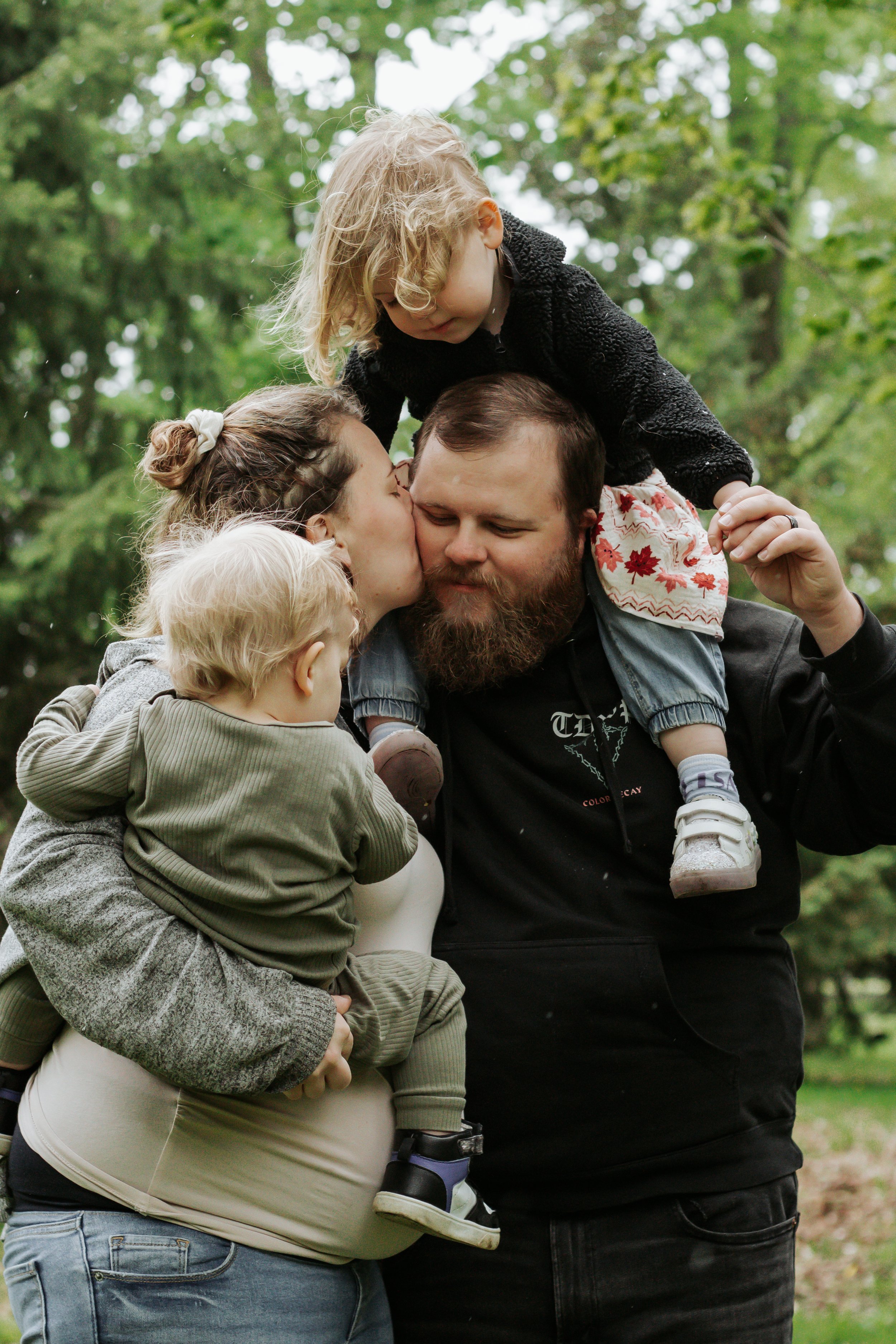 A family of five outdoors hugging and kissing on a cloudy day surrounded by green trees. Family photos at elgin park uxbridge ontario