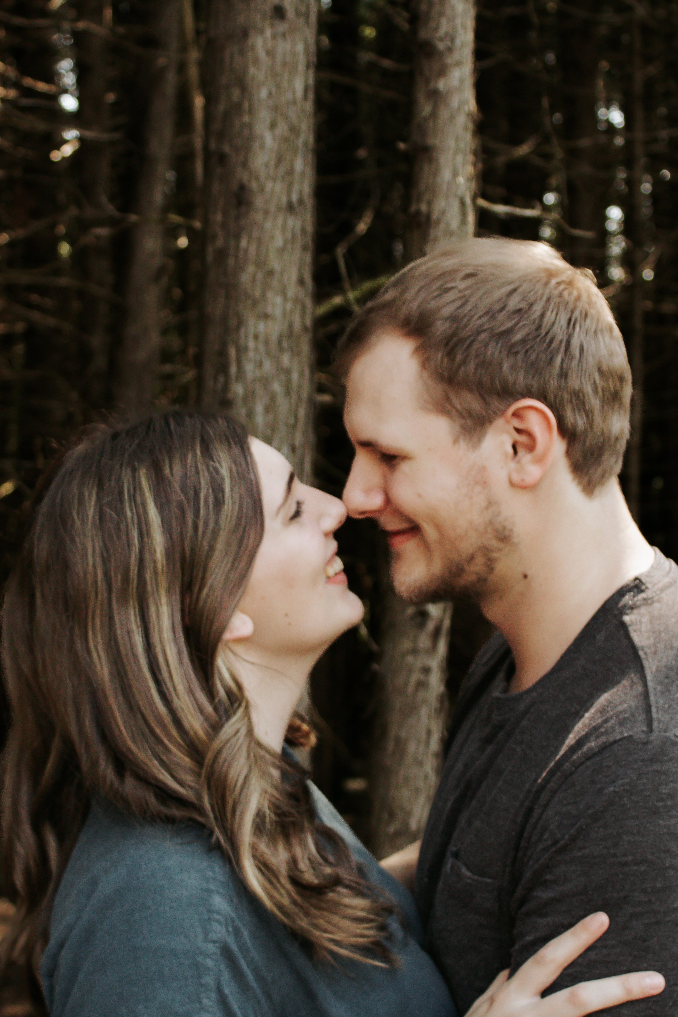 A young couple standing close together, leaning in for a kiss, outdoors with trees in the background.