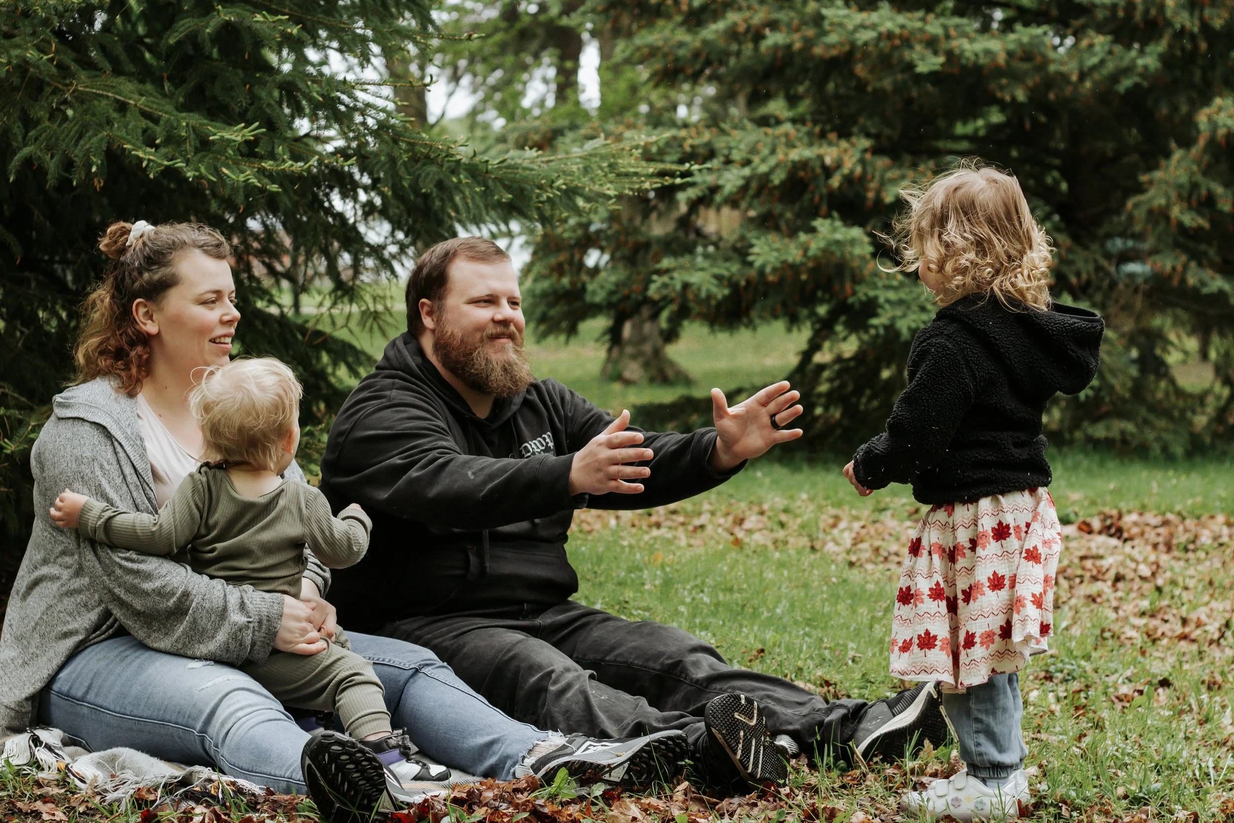 A family of four sitting on the grass in a park surrounded by trees. The mother holds a small child, the father is reaching out to a young girl standing in front of them. Family photos at elgin park uxbridge ontario