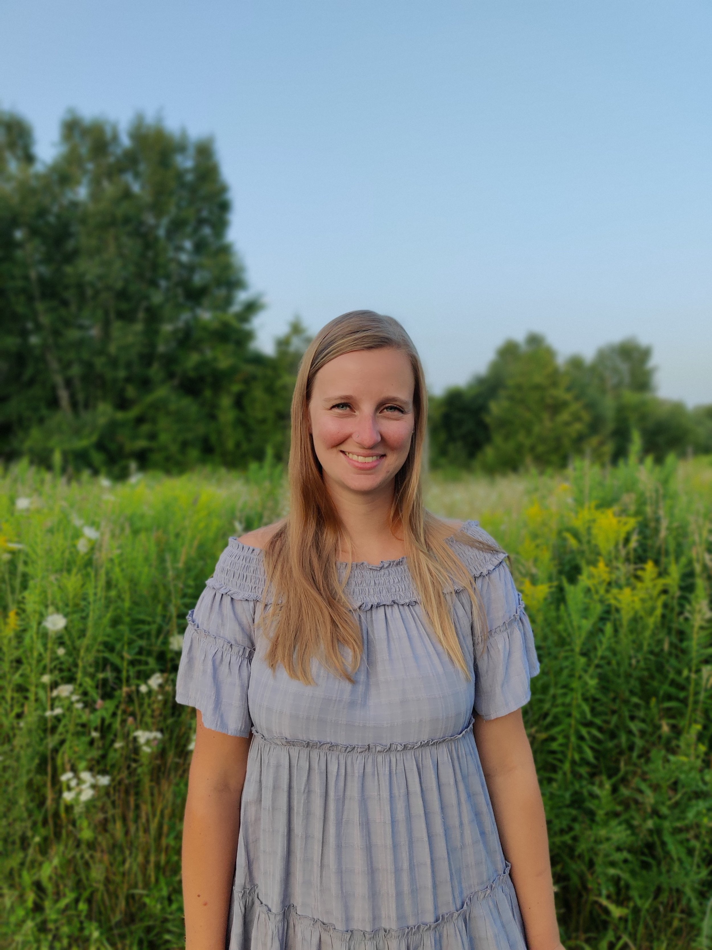 A young woman with long blonde hair smiling outdoors in a green field with trees in the background, during daytime.