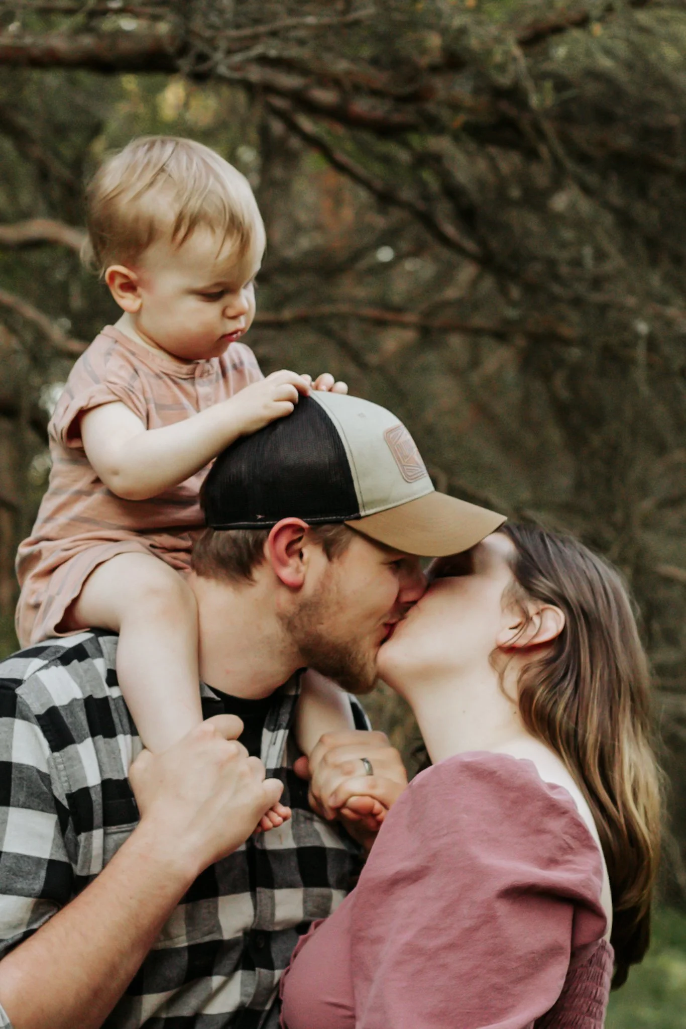 A young child sitting on a man's shoulders, both of them kissing a woman. The scene appears to be outdoors with trees in the background. maternity photos Uxbridge ontario