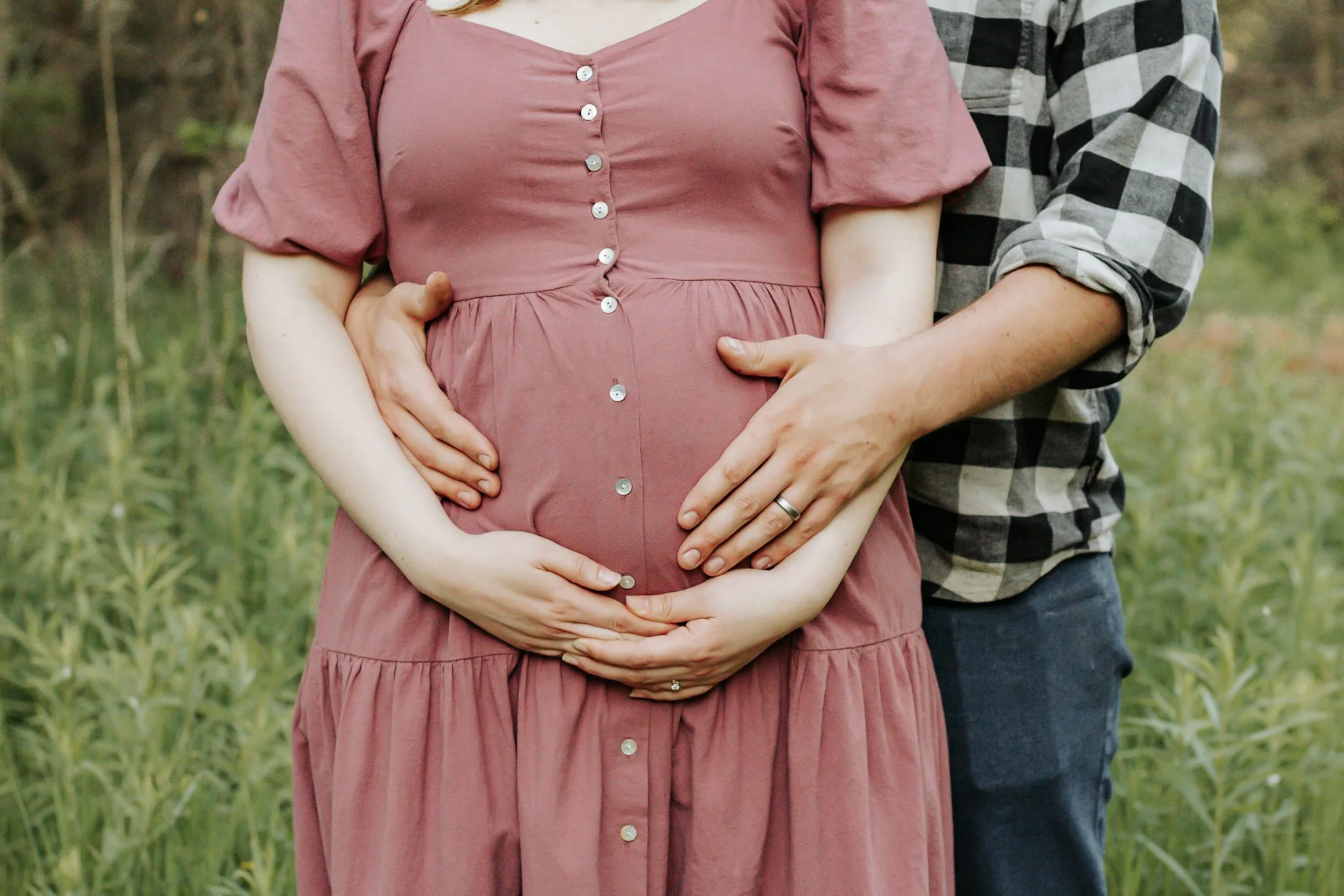 maternity photos Uxbridge ontario A pregnant woman in a mauve dress standing outdoors, with a man beside her. Both have their hands on her belly, with the man's hand over hers. The woman is wearing a ring, and the background shows green grass and tre