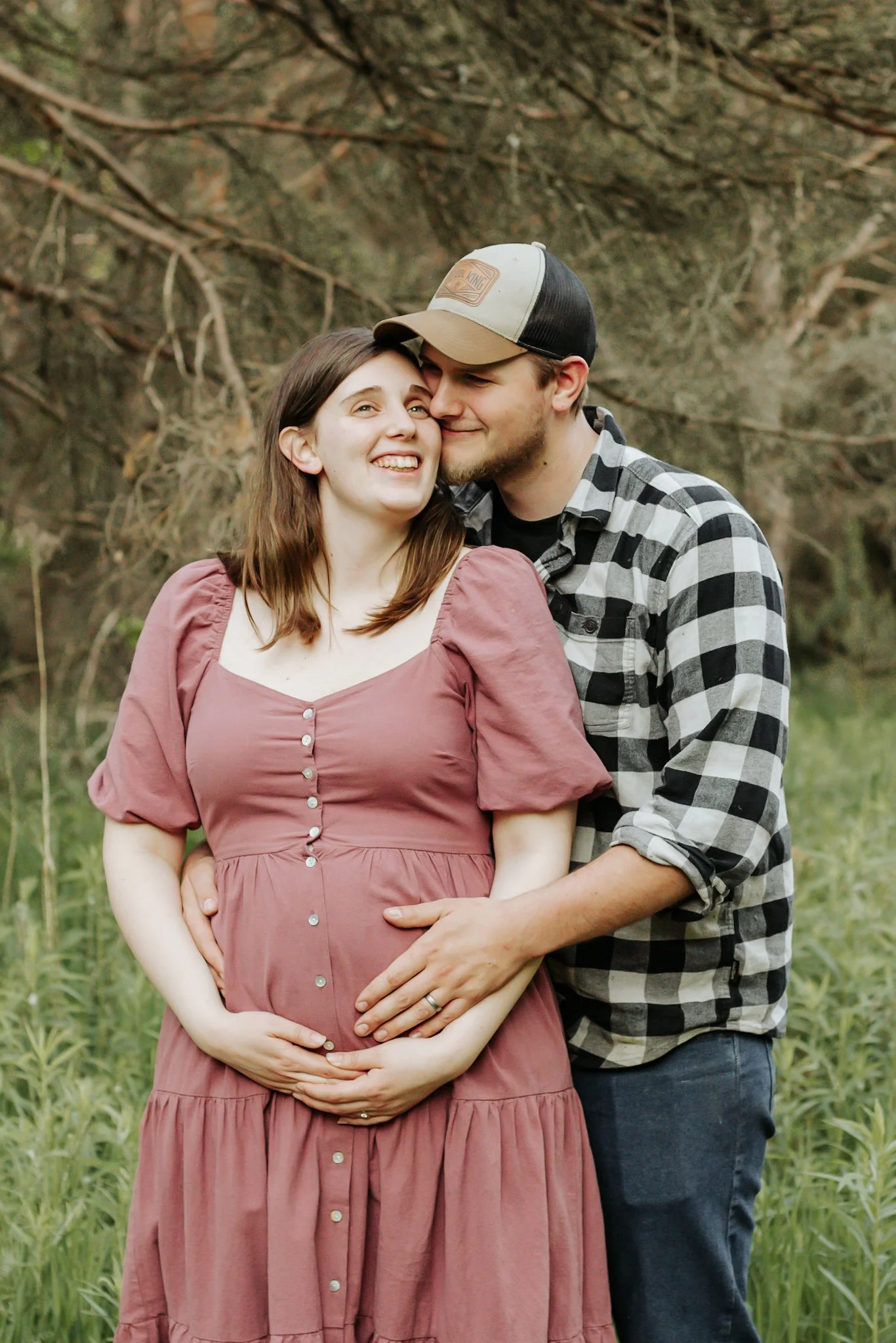 A couple, with the man embracing the pregnant woman, standing outdoors in a grassy area with trees in the background. The woman is smiling and holding her belly, while the man is leaning in close and smiling. maternity photos Uxbridge ontario