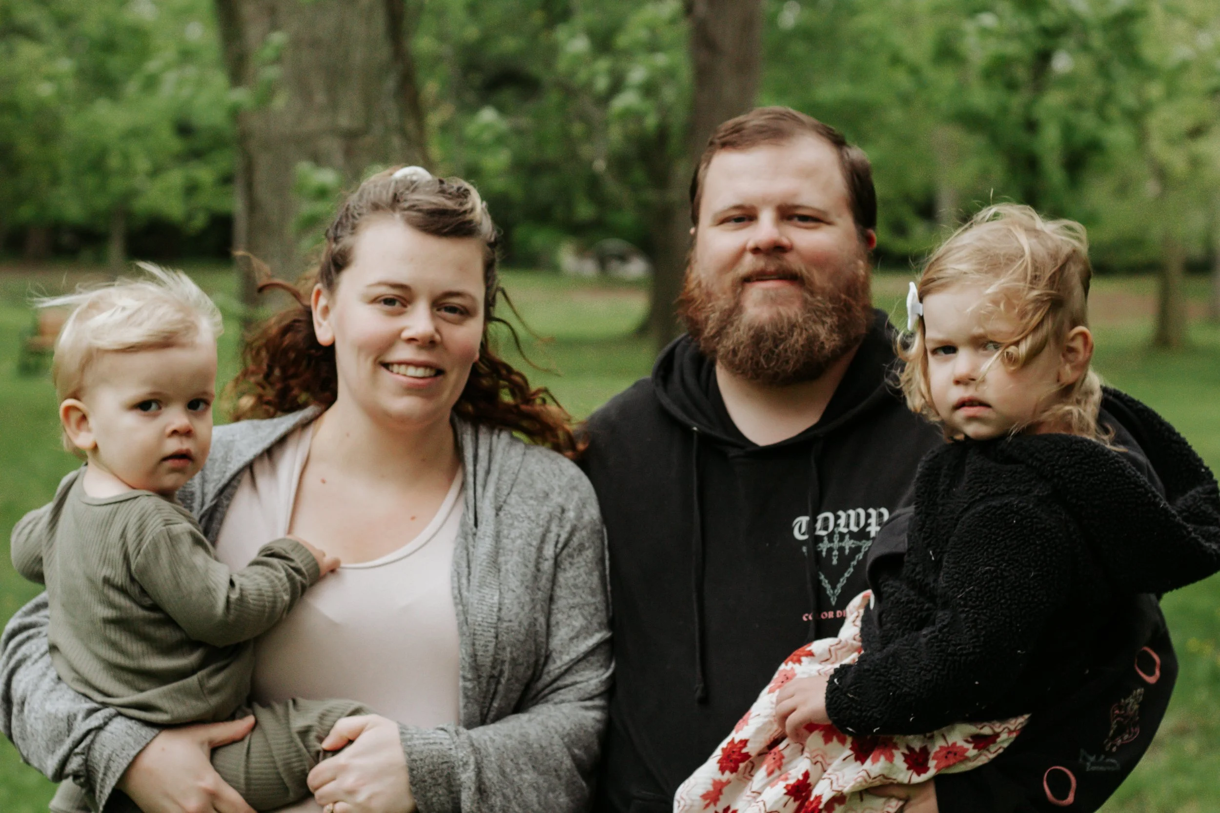 A family of four stands outdoors in a park with trees in the background. The woman has curly brown hair and is smiling, holding a young child with blonde hair and a serious expression. The man has a beard and is smiling, holding a young girl with blo