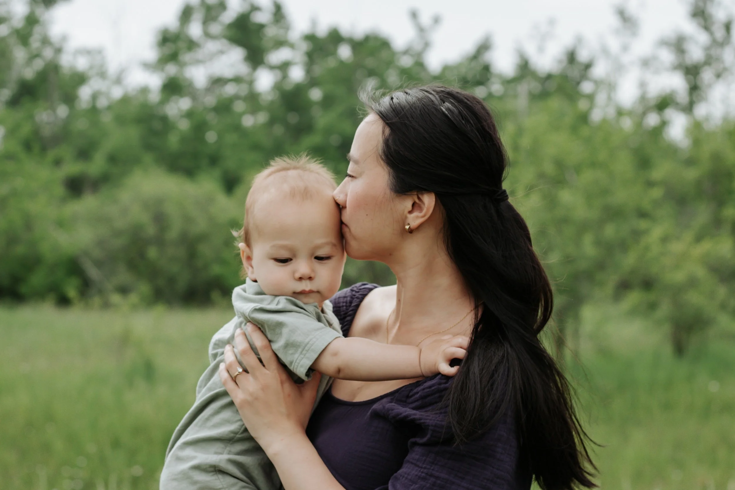 A woman holding a toddler in an outdoor setting with green trees and grass, woman is kissing the toddler's forehead.
