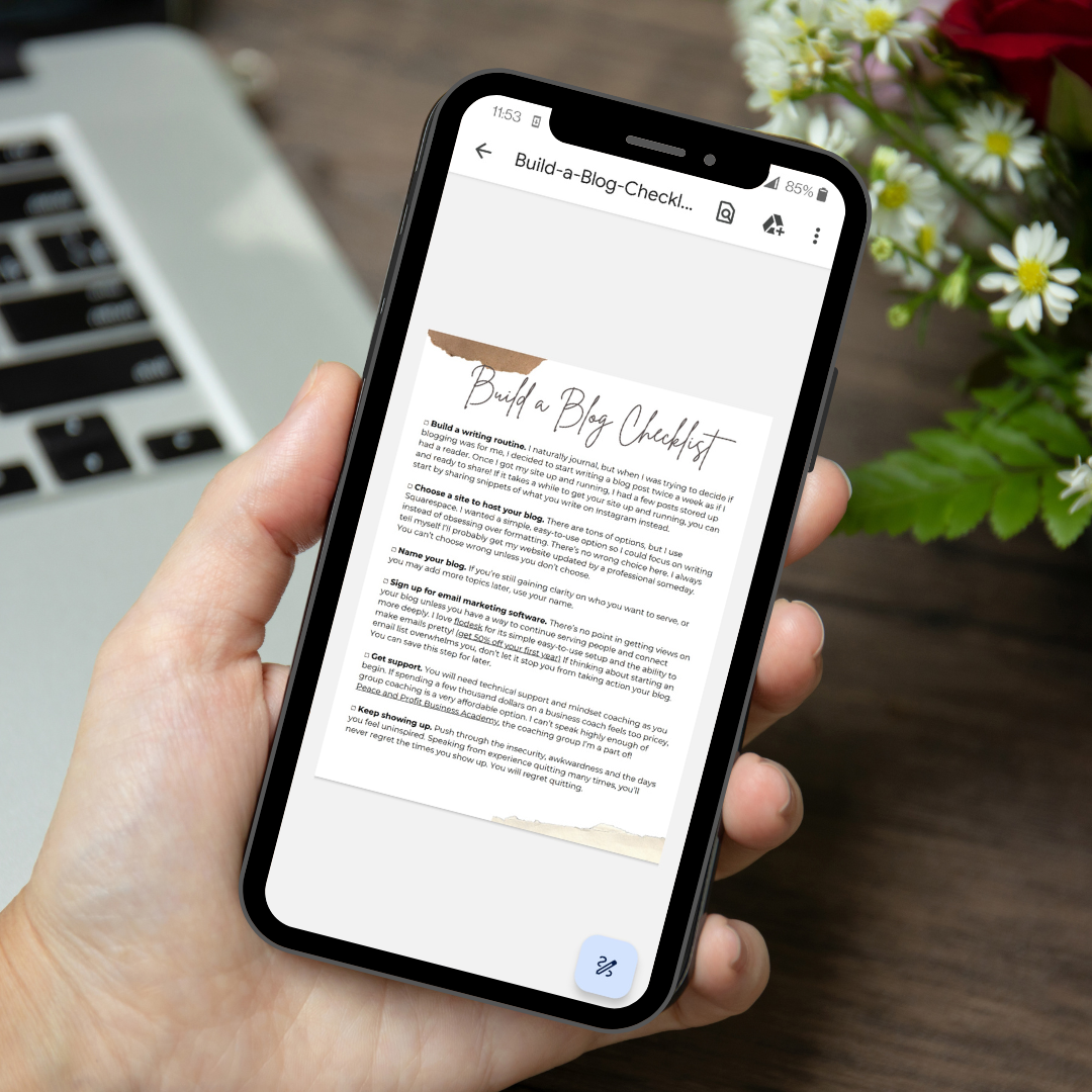 A person holding a smartphone displaying a blog checklist titled 'Build a Blog Checklist' with a flower arrangement and keyboard in the background.