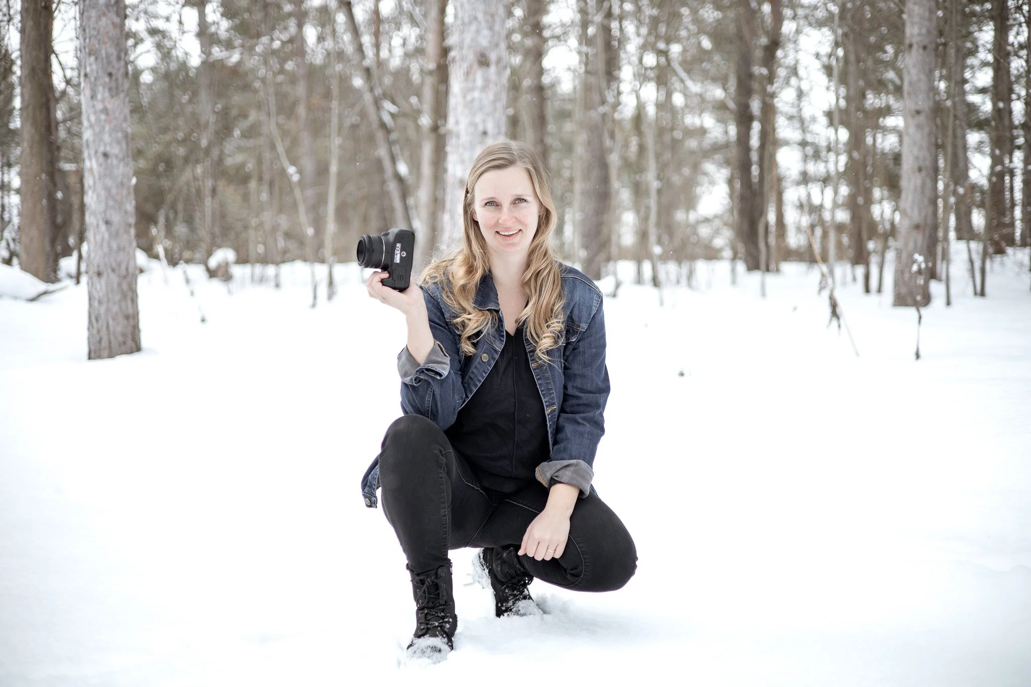 Uxbridge family photographer, Micaela Bouwmeister of Micaela B Photography poses kneeling in the snow in a wooded area, holding a camera in her right hand, smiling at the camera.