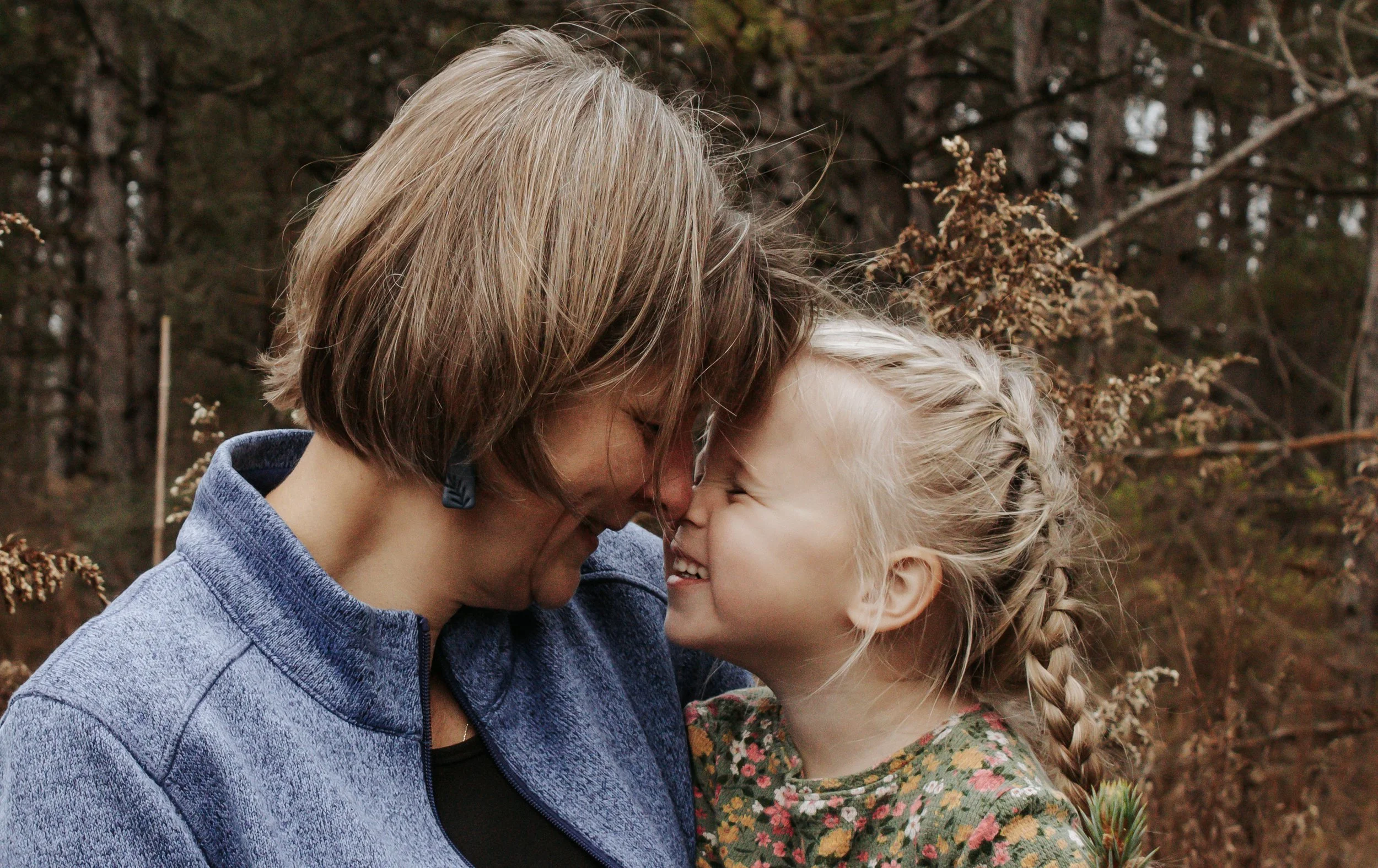 A woman and a young girl are touching foreheads and smiling in a wooded outdoor setting.