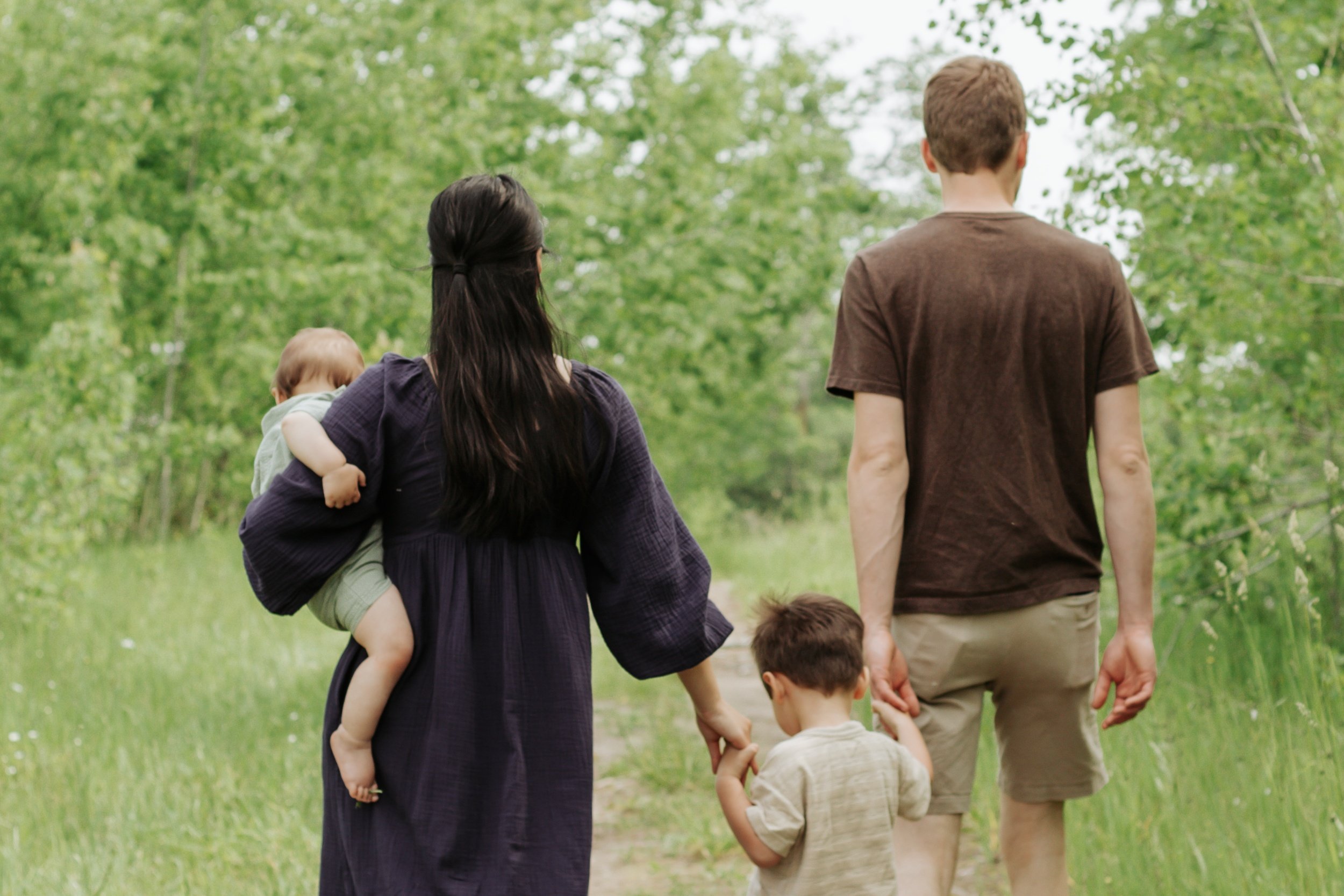 A family of four, including two children, walks along a wooded trail holding hands during daytime.