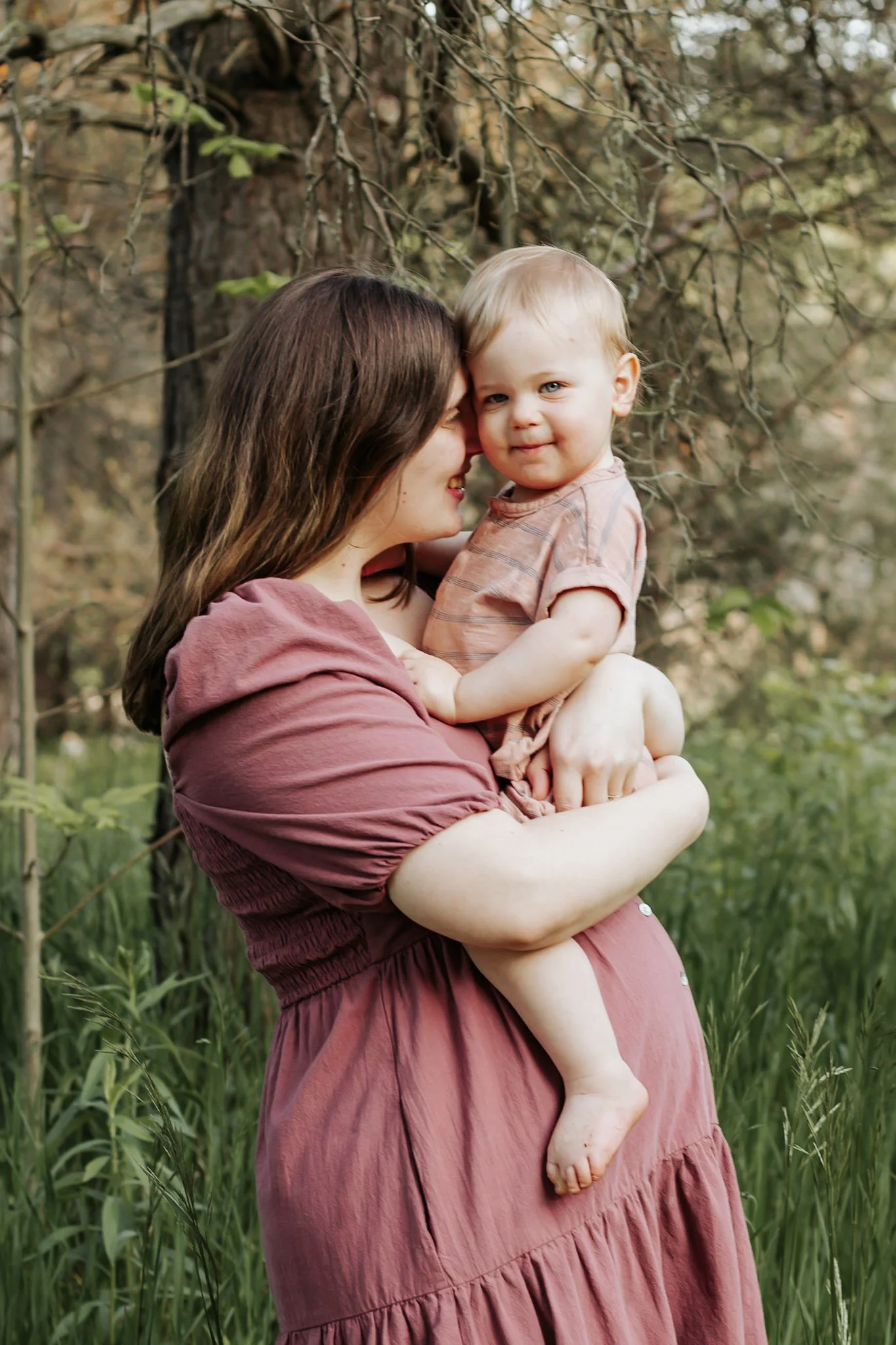 A woman with brown hair in a mauve dress holding a young blond-haired child outside in a wooded area. maternity photos Uxbridge ontario