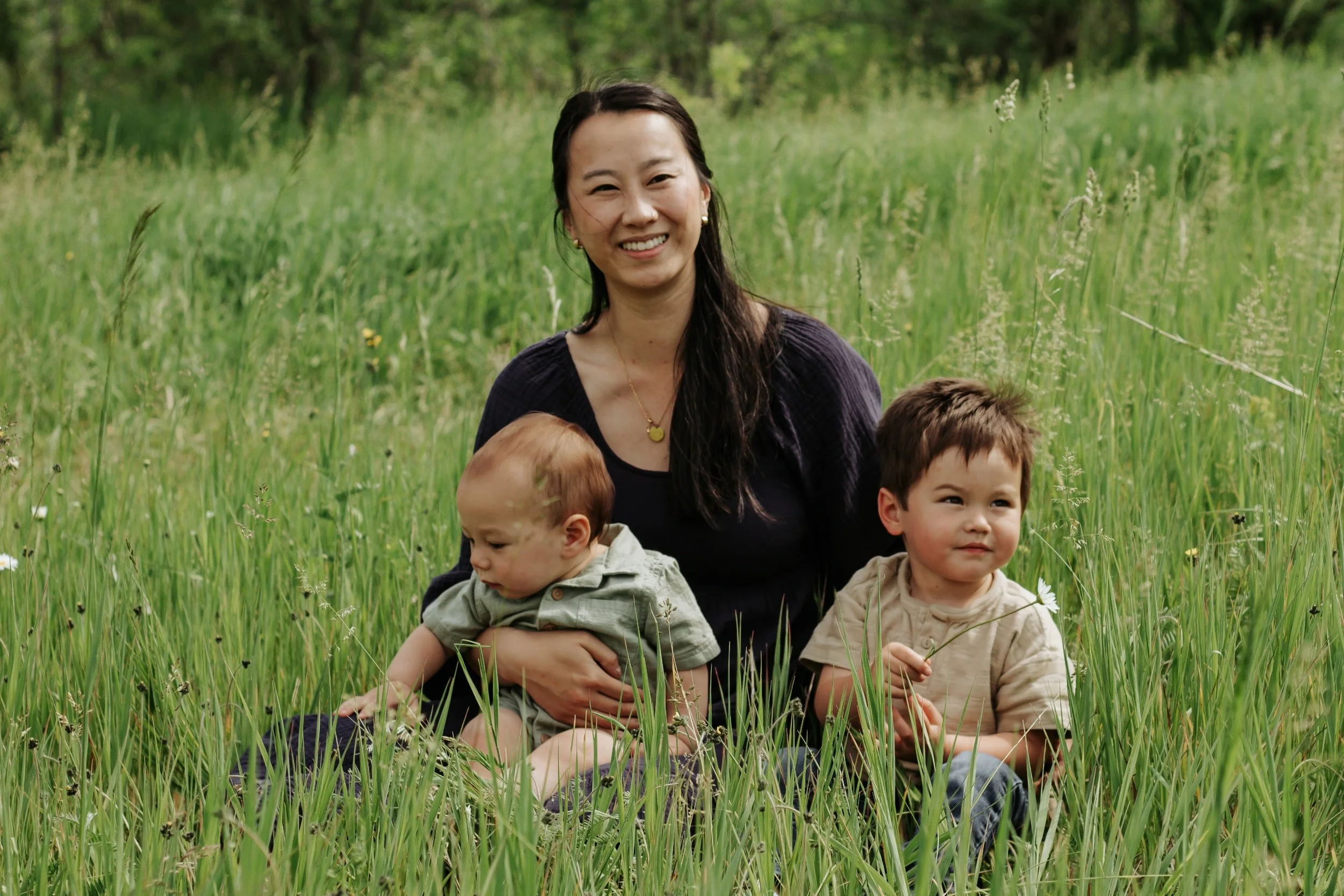 A woman sitting in a field with two young children, one child sitting on her lap and the other sitting beside her, all surrounded by green grass and wildflowers. Family photos in sunderland ontario