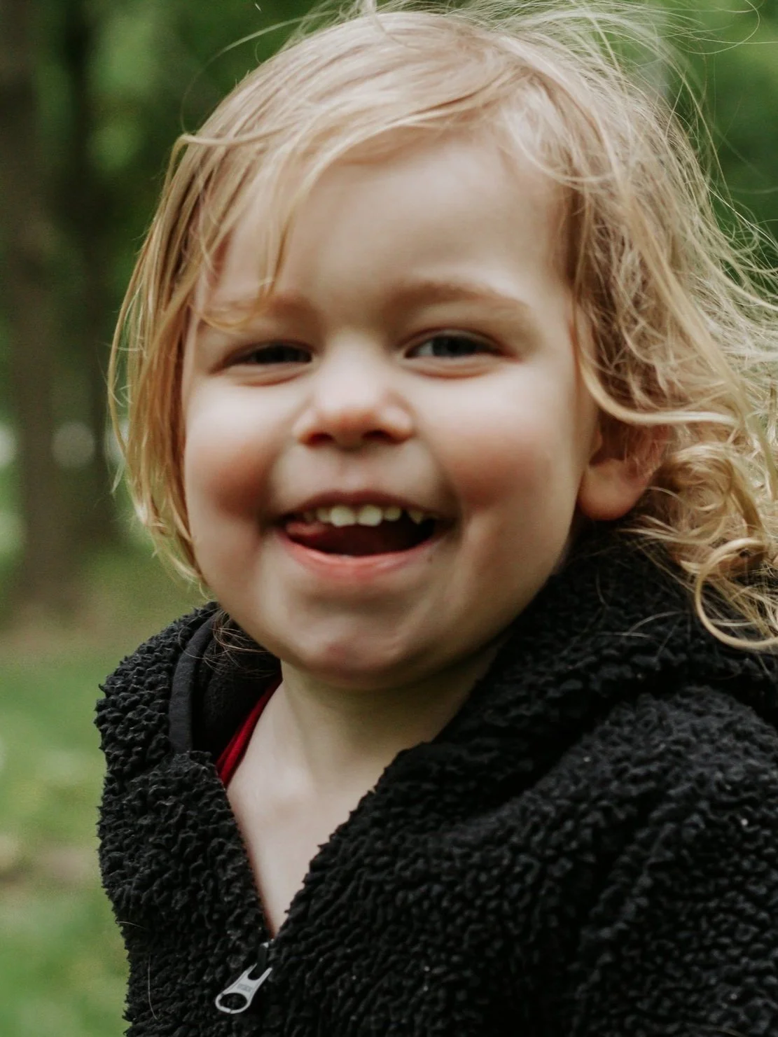 A young child with curly blonde hair smiling outdoors in a natural setting.