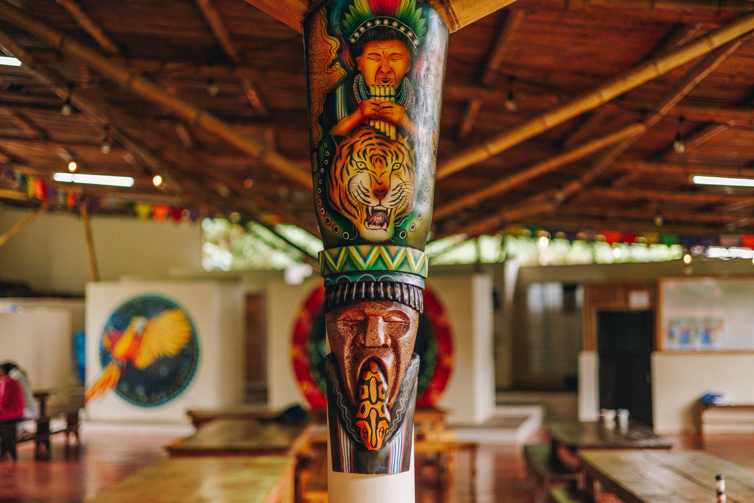 Decorative totem pole with a painted figure of an indigenous person playing a flute and a tiger's face and tongue below, inside a wooden pavilion with tables and colorful art on the walls.
