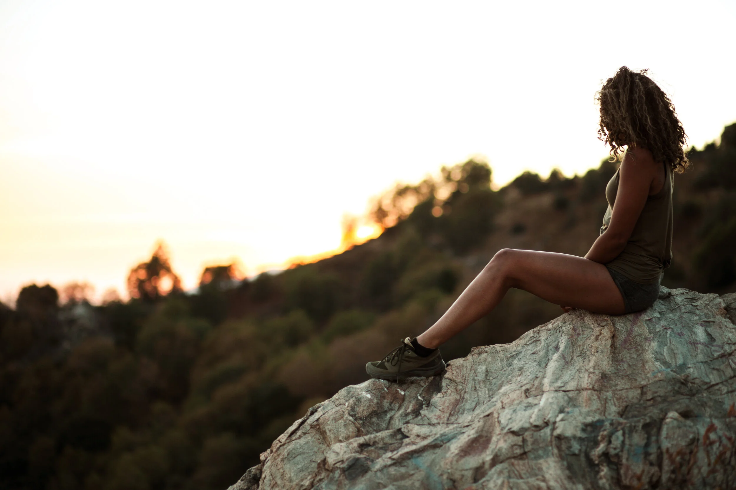A woman sitting on a rocky ledge during sunset in a mountainous area.
