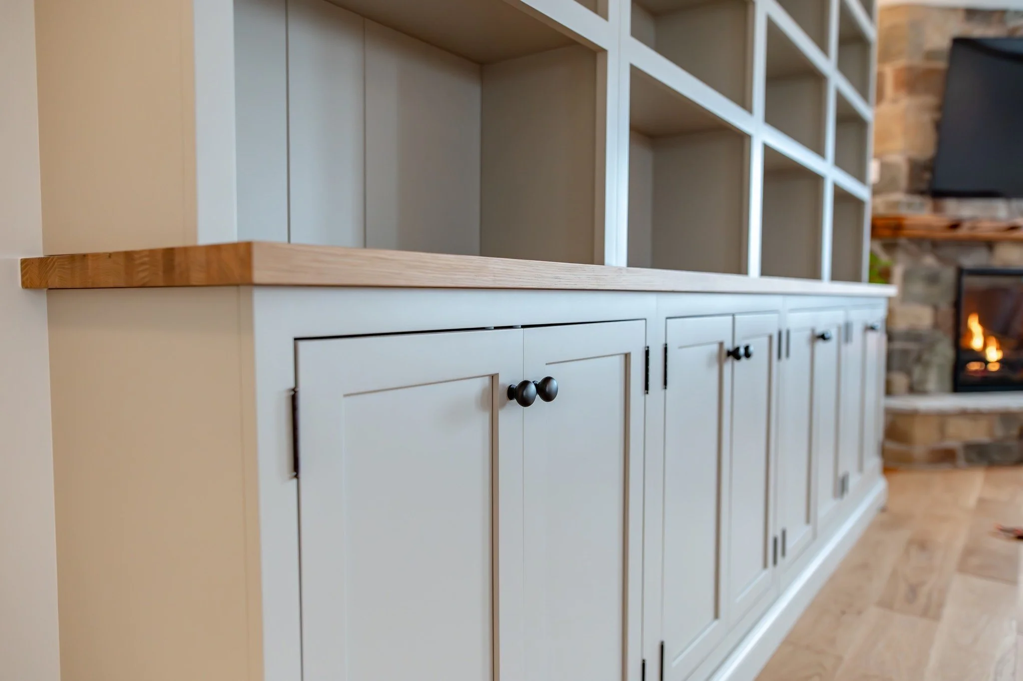 Modern country style built-in bookcase, greige lower cabinets with open shelving above with v-groove back panels, dark bronze hardware, next to fieldstone fireplace with reclaimed wood beam mantle