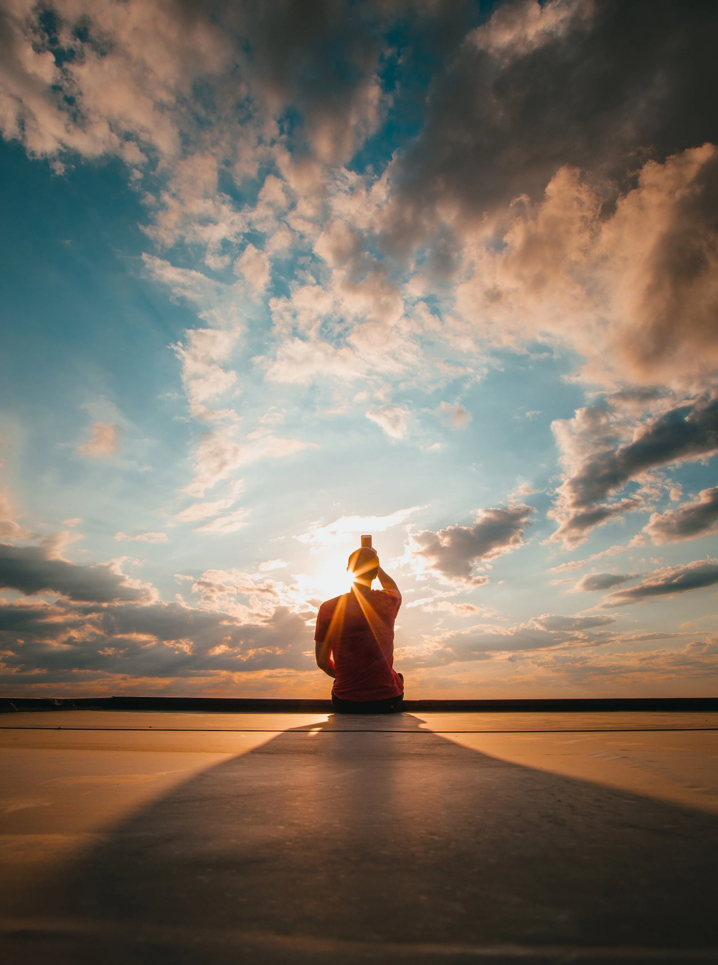 Person sitting on a ledge outdoors during sunset or sunrise, taking a photo with their phone, with the sun behind them and clouds in the sky.