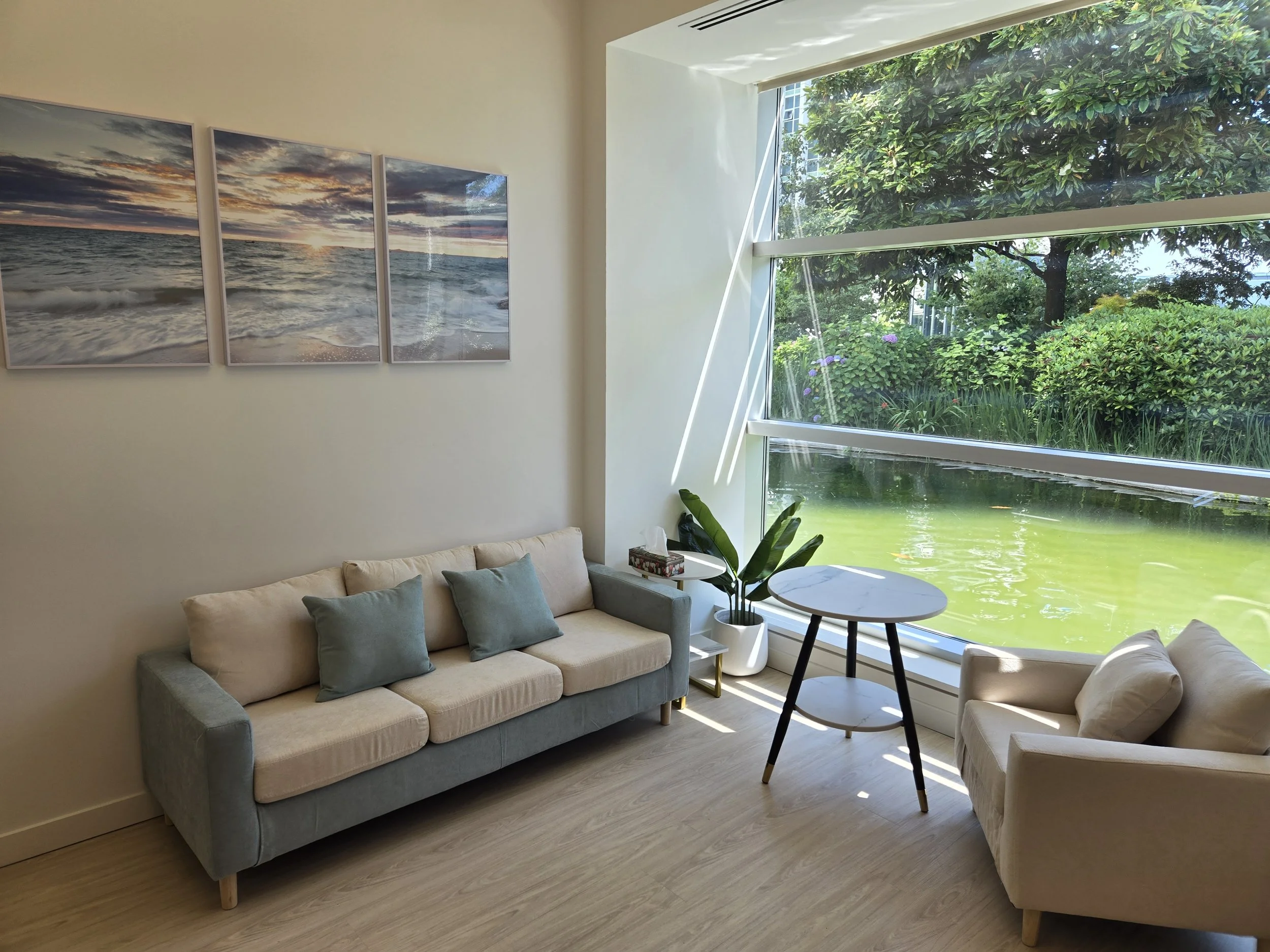Counselling room in Vancouver with a large window overlooking a pond and garden, beige sofa with blue cushions, small round table, armchair, potted plant, and wall art depicting a sunset over the ocean.