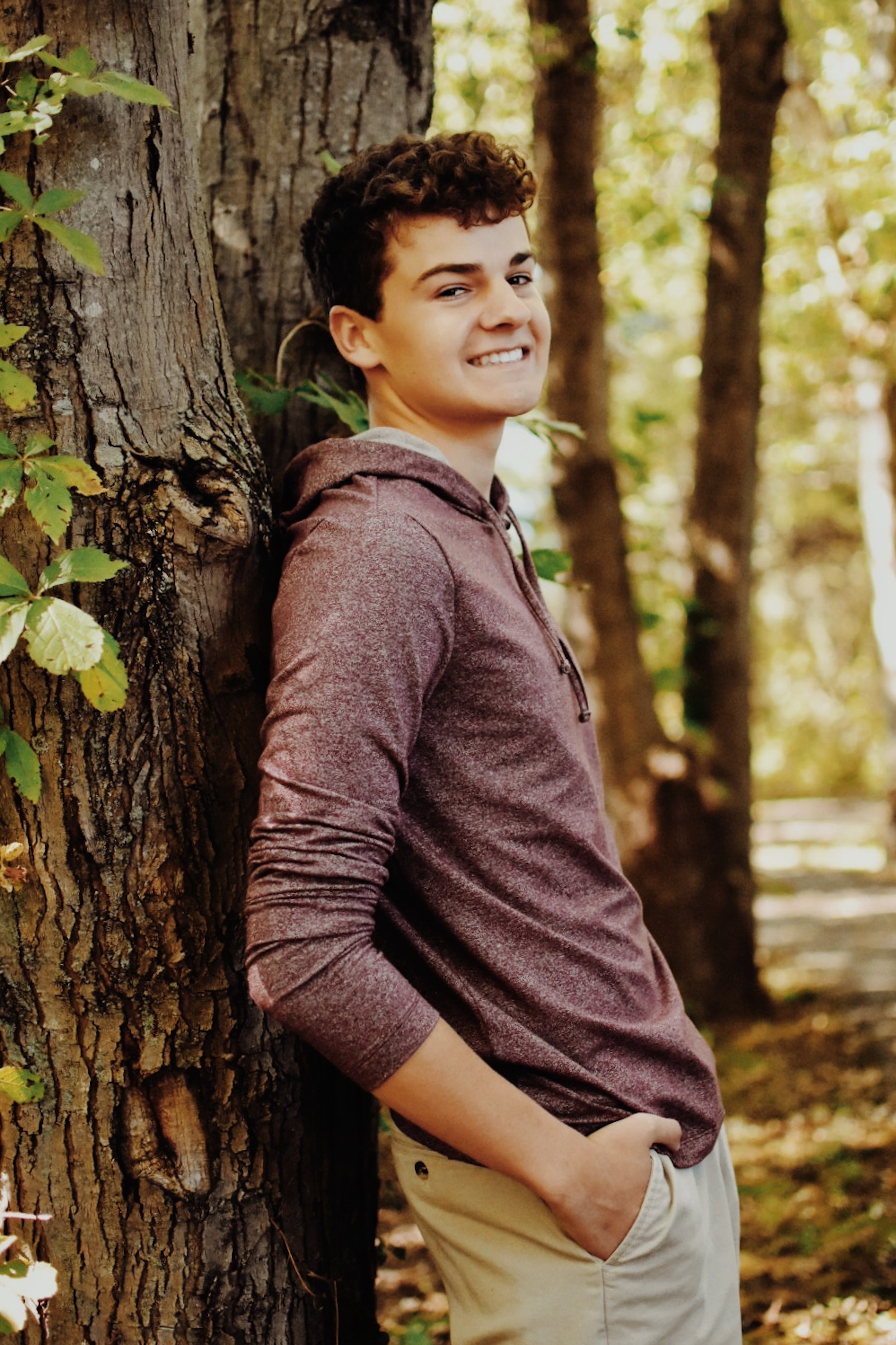 A teenage boy with curly hair standing next to a tree, smiling, with his hands in his pockets, in a wooded area during daytime.