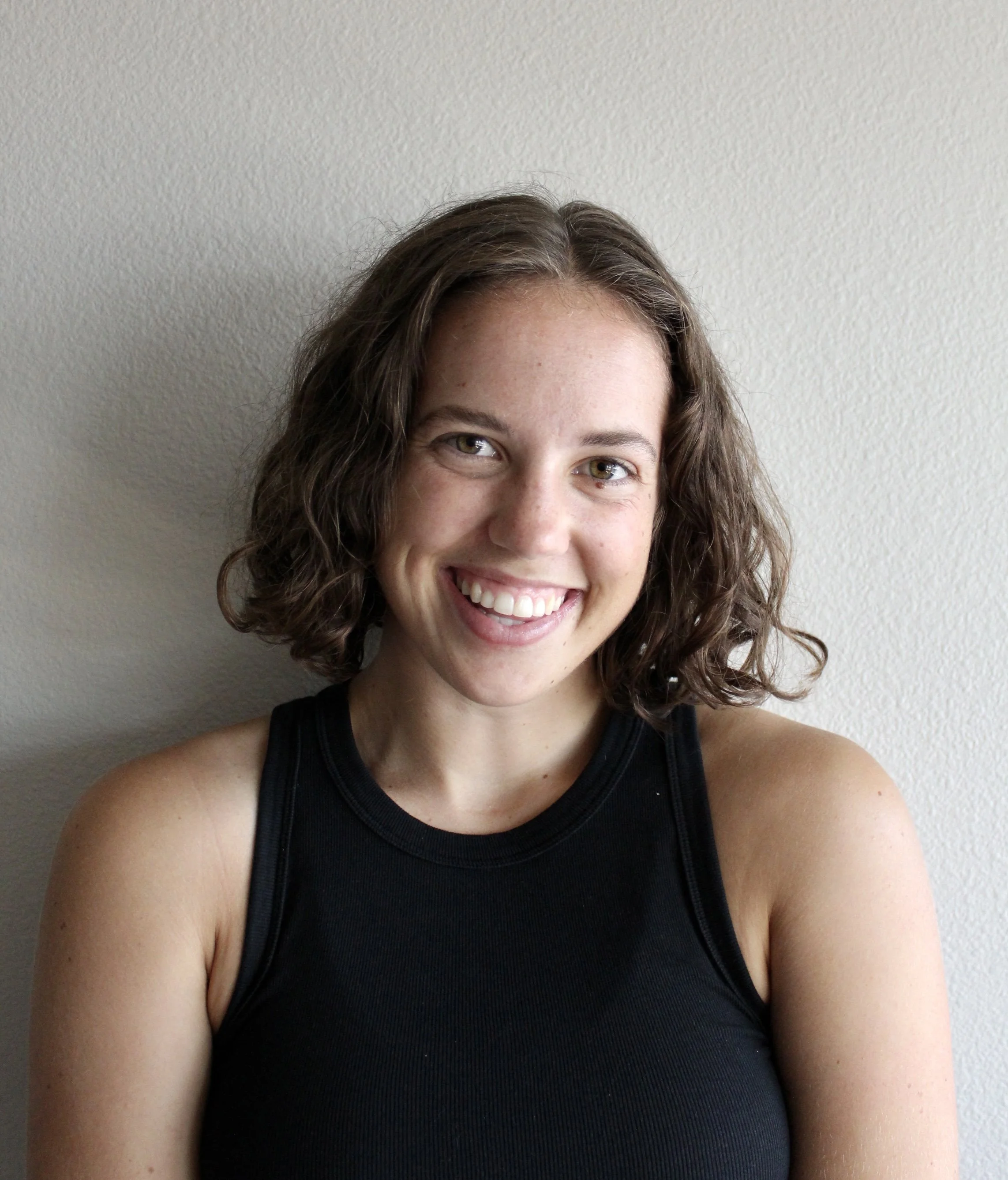 Close-up of a woman smiling, with brown wavy hair and wearing a black sleeveless top, standing against a plain light-colored wall.