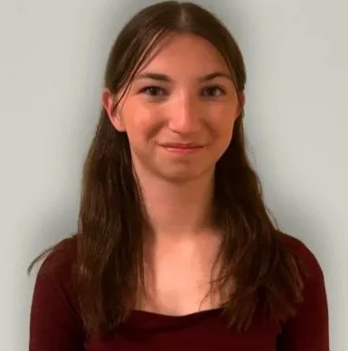 Young woman with long brown hair and a slight smile, wearing a maroon top, standing against a light green background.