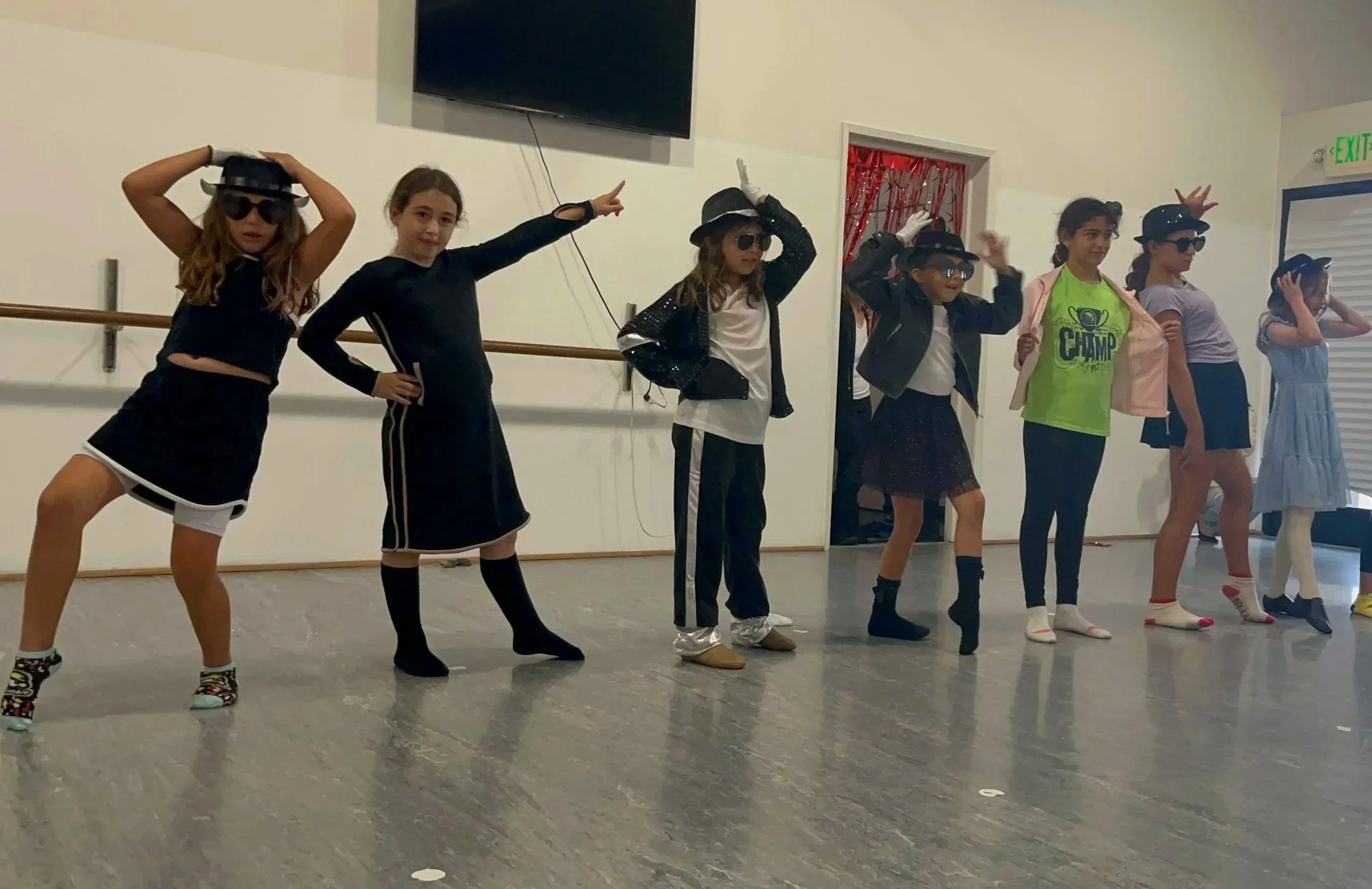 A group of children standing in a dance studio, dressed in casual and dance costumes, some with sunglasses and hats, posing in a line for a dance or performance.