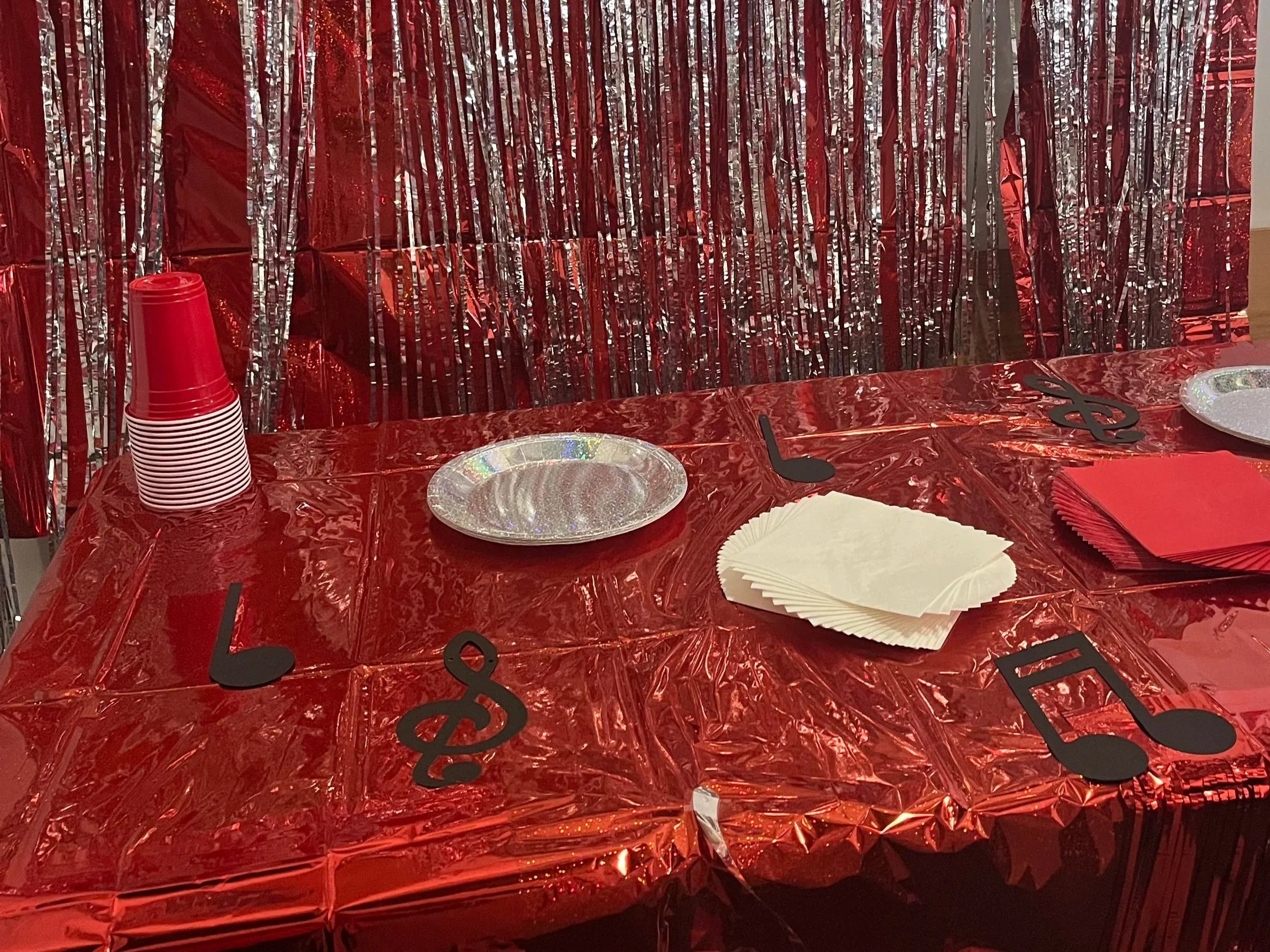 A table decorated for a celebration with a red metallic tablecloth, black cutout letters spelling 'Love', stacks of red plastic cups, paper plates, napkins, and a metallic red tinsel backdrop.