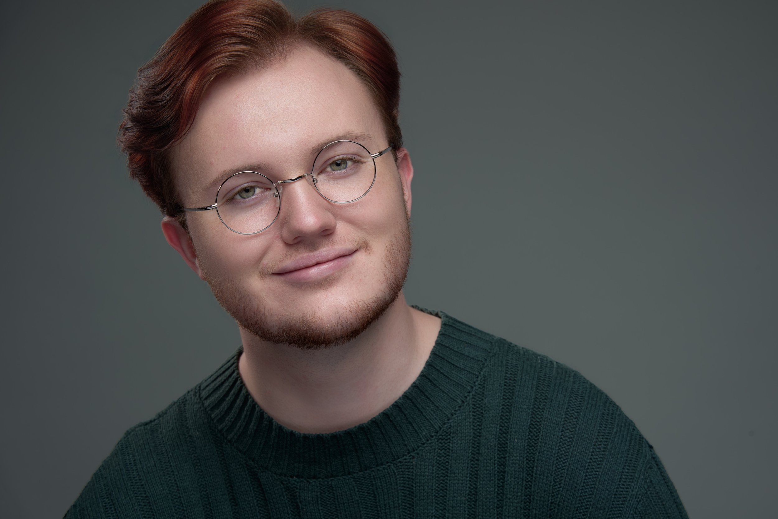 A young man with red hair, glasses, and a beard smiling, wearing a dark green sweater against a plain gray background.