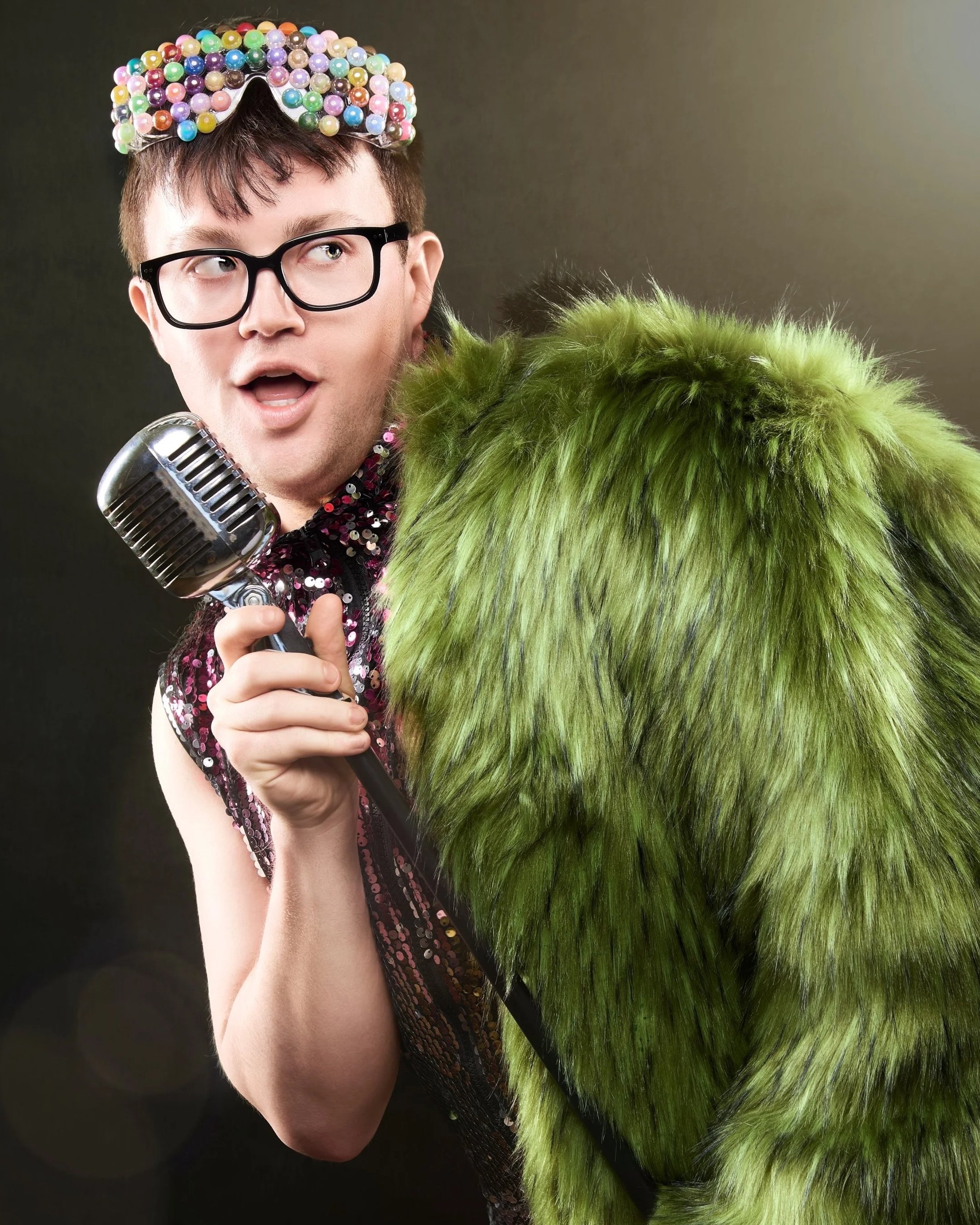 A man with glasses wearing a colorful, beaded crown, holding a vintage microphone, and dressed in a sequin top, standing next to a large, furry, green costume or prop.