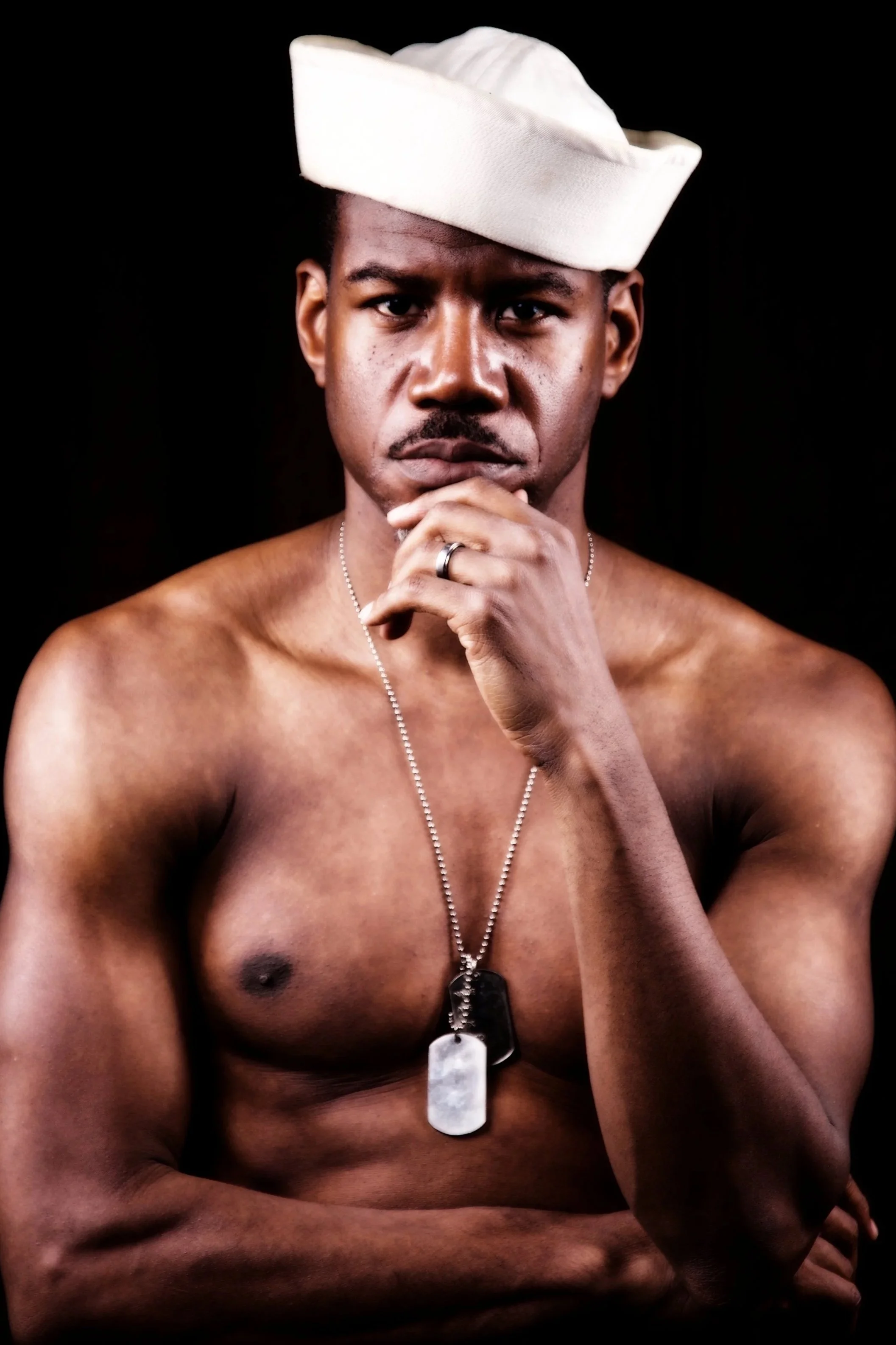 A shirtless man with dark skin, short hair, and a serious expression, wearing a white sailor hat and dog tags around his neck, against a black background.