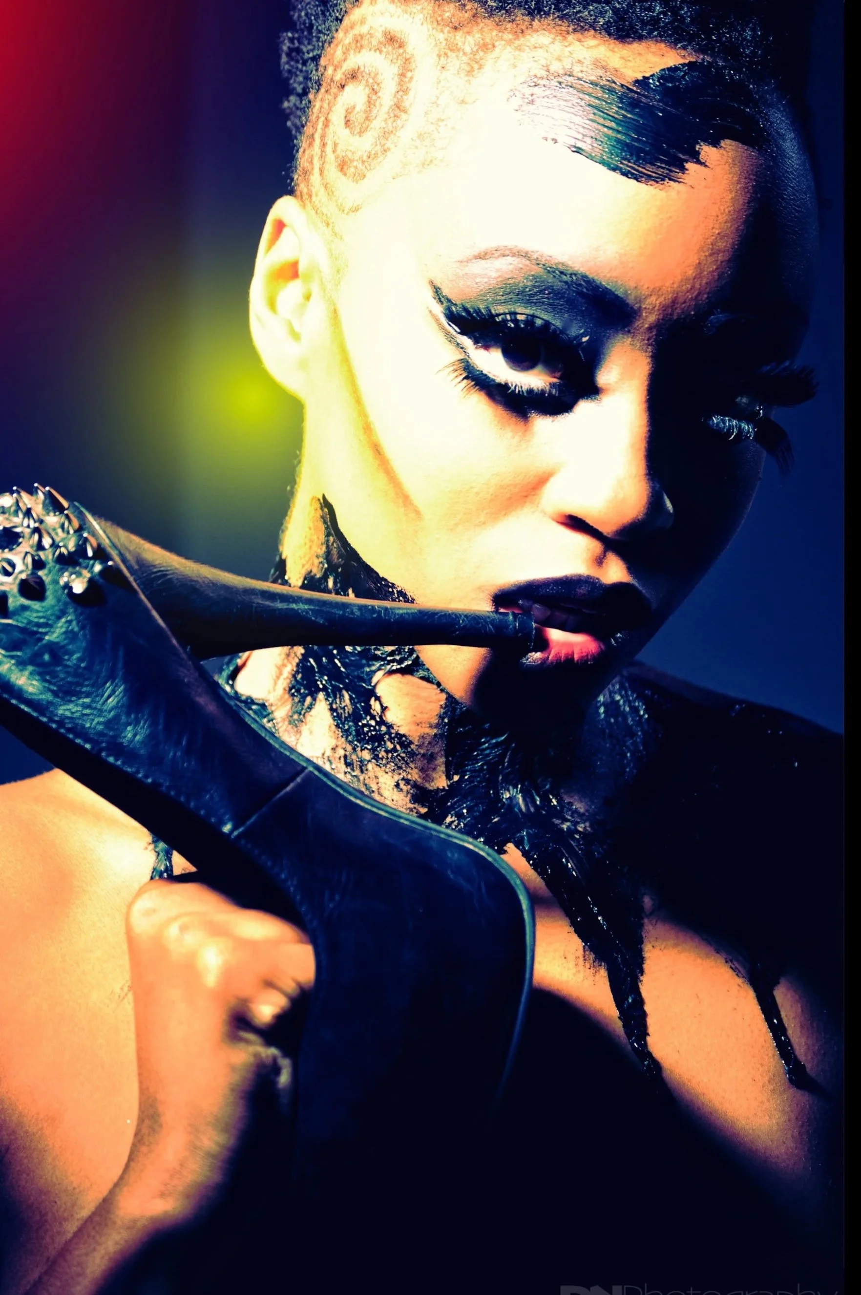 A woman with an edgy hairstyle; shaved sides with a spiral design, and styled hair on top. She has bold makeup with dark eyeshadow, thick eyeliner, and dark lipstick. She is holding a high-heeled shoe with metallic spikes near her face, and her body 