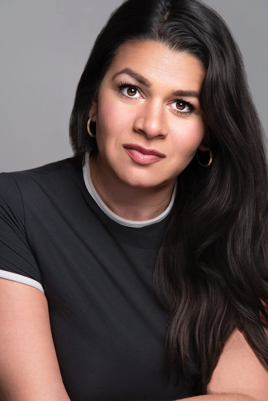 A portrait of a woman with long dark hair, wearing hoop earrings and a black shirt with white collar trim, against a gray background. Darius Nichols Photography DNPhotographyNYC