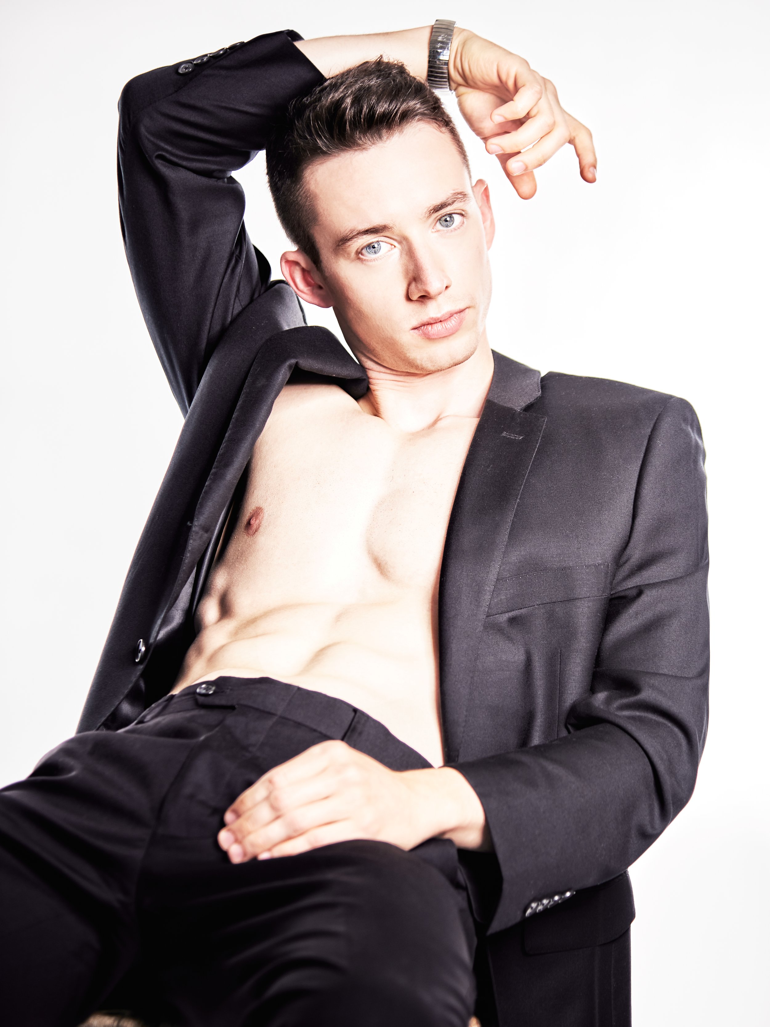 A young man with short dark hair, wearing a black blazer, is lifting his blazer to reveal his bare chest and abs, posing against a plain white background.