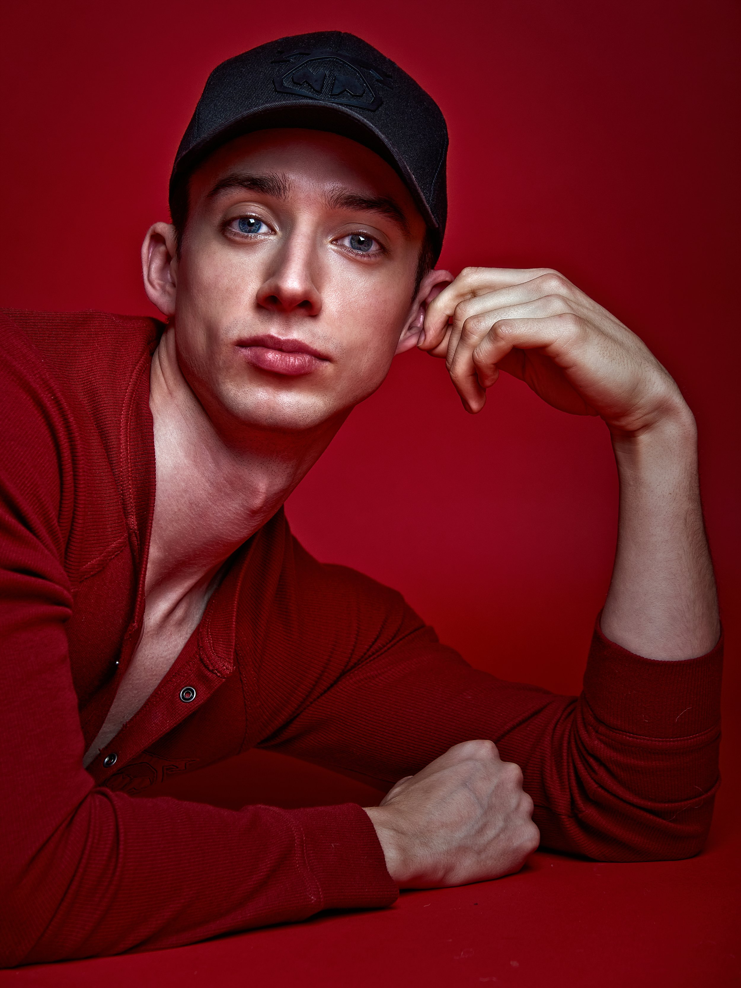 Young man with blue eyes wearing a black cap, lying on a red surface with a red background, looking at the camera. Darius Nichols Photography DNPhotographyNYC