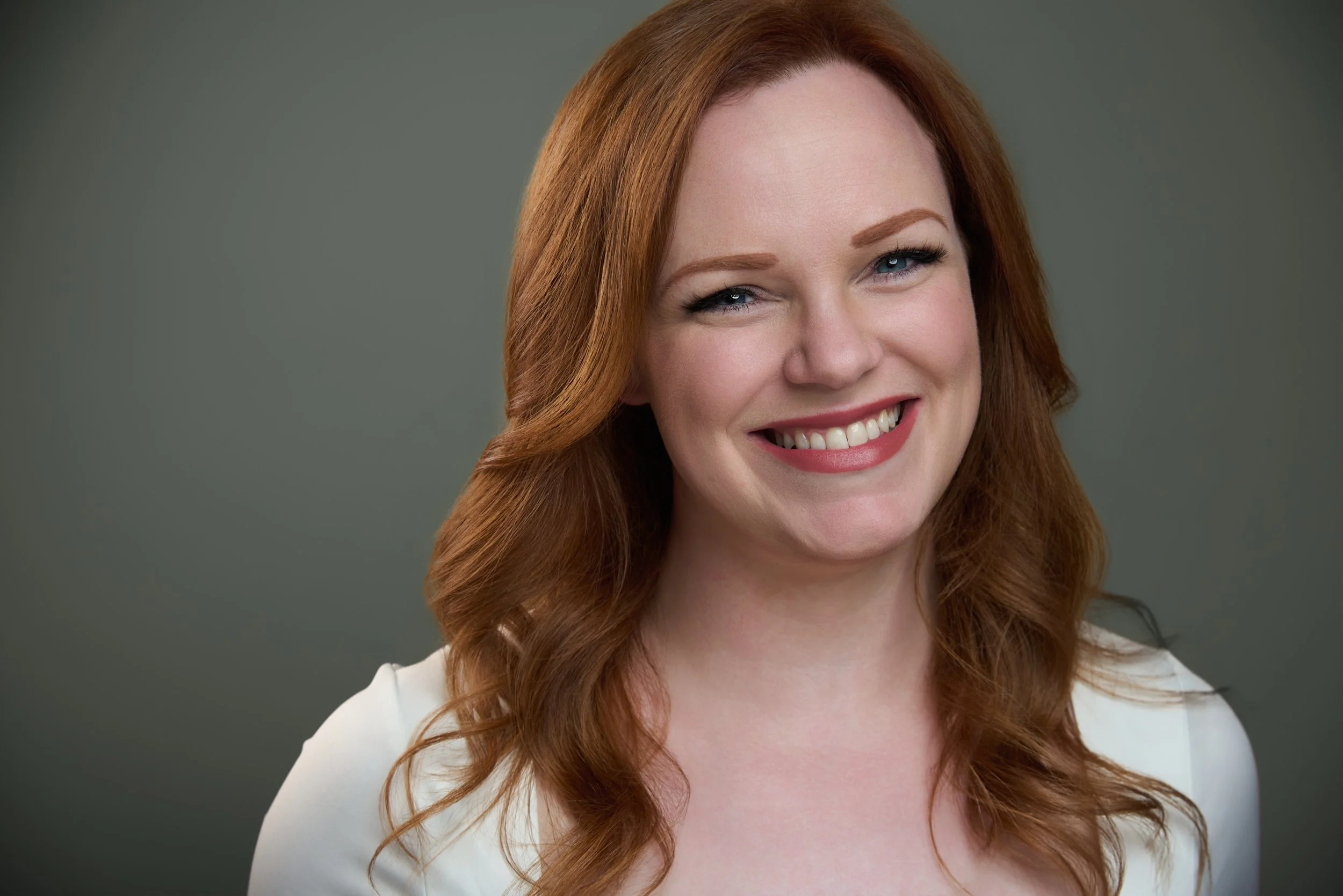 A smiling woman with long, wavy red hair and fair skin, wearing a white top, against a plain gray background.