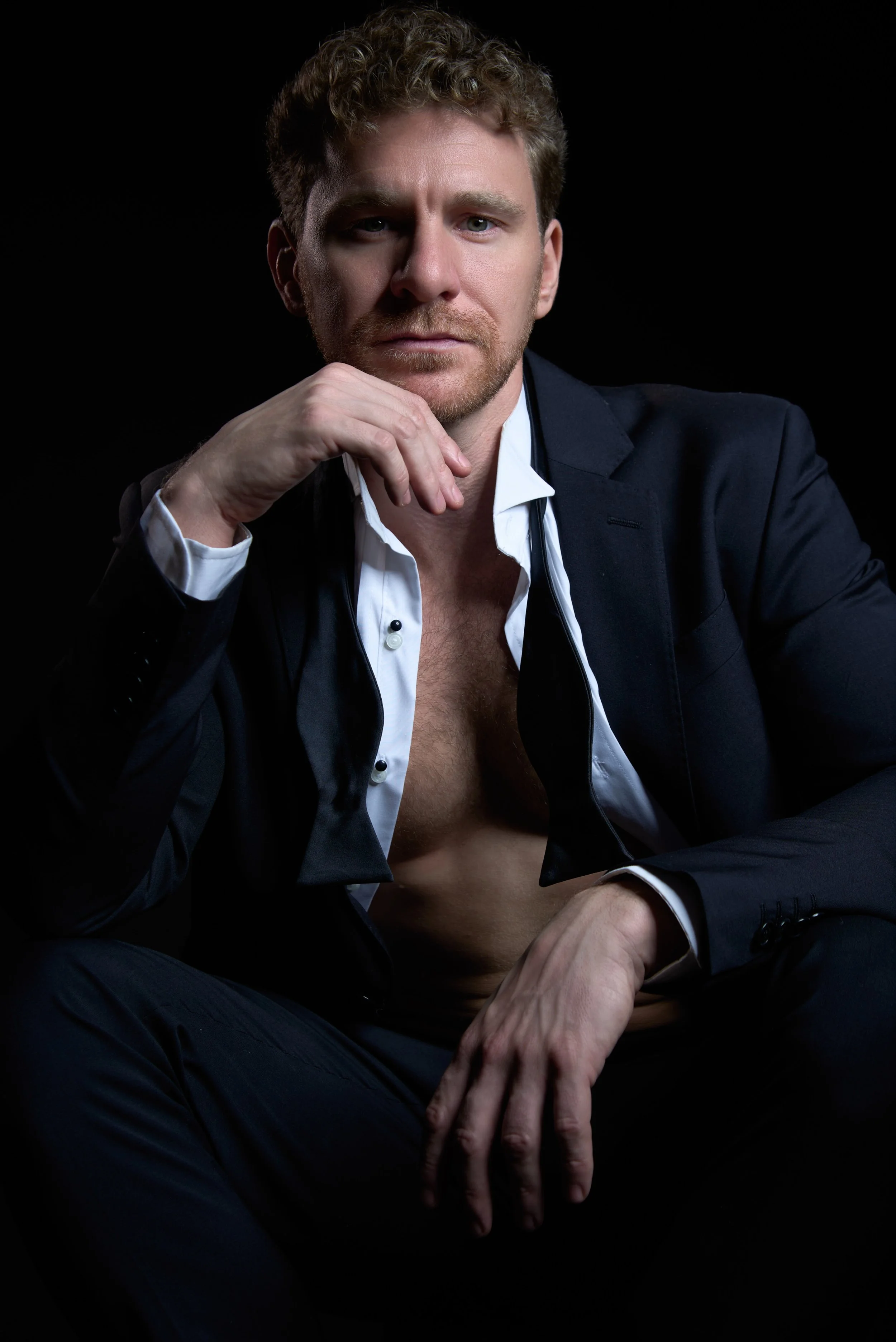 A man with curly hair and a beard wearing a dark suit jacket with a white shirt, sitting against a black background. His shirt is partly unbuttoned, exposing his chest, and he is resting his chin on his left hand with a serious expression.
