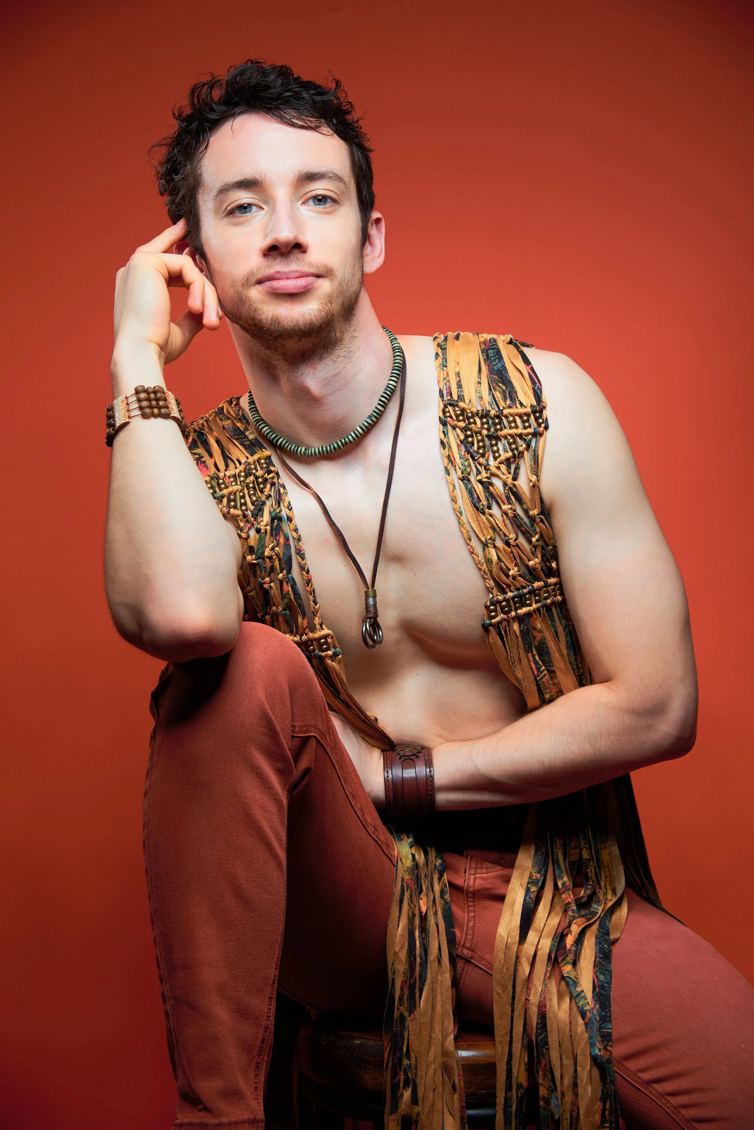 A young man with dark, curly hair and a light beard poses confidently against a warm, orange background. He is shirtless, wearing a patterned vest, brown pants, and various accessories including a beaded necklace, a leather wristband, and multiple br